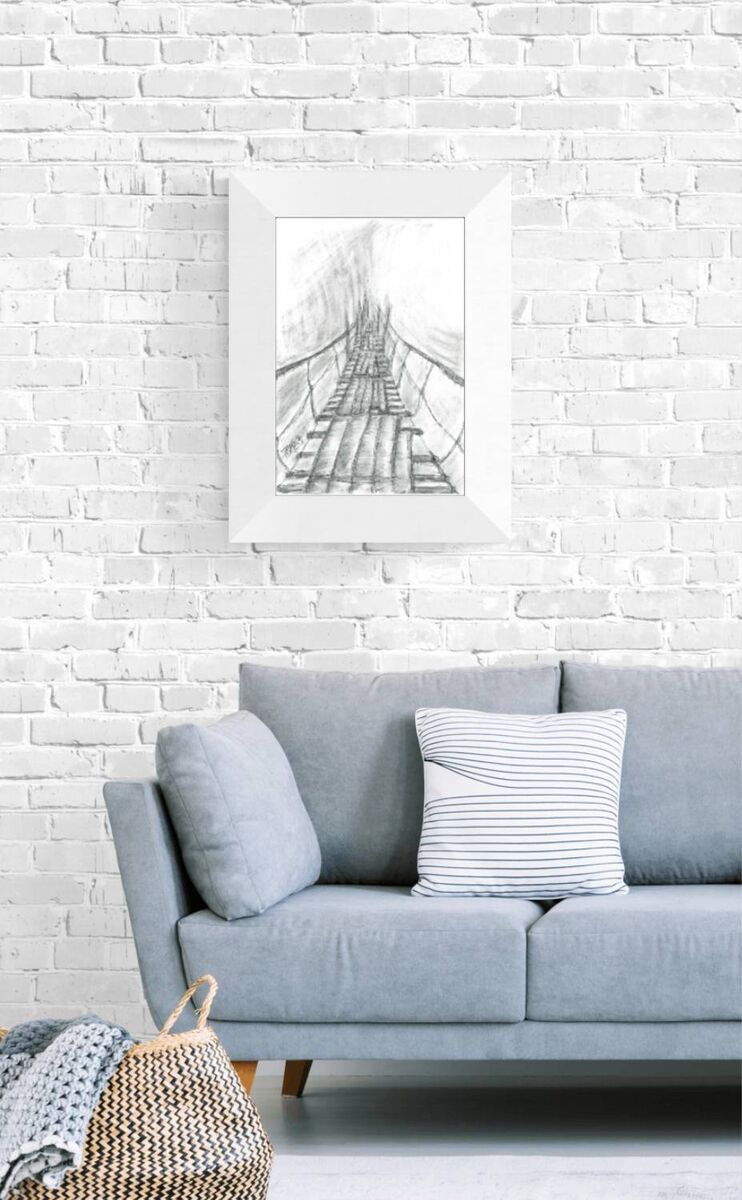 Original surreal charcoal drawing Toward the Unknown by Diana Dimova TRAXI, featuring a suspension bridge in mist exploring transition in black and white. A framed black and white sketch of a suspension bridge hanging over water, displayed on a white brick wall above a light blue sofa with decorative pillows.