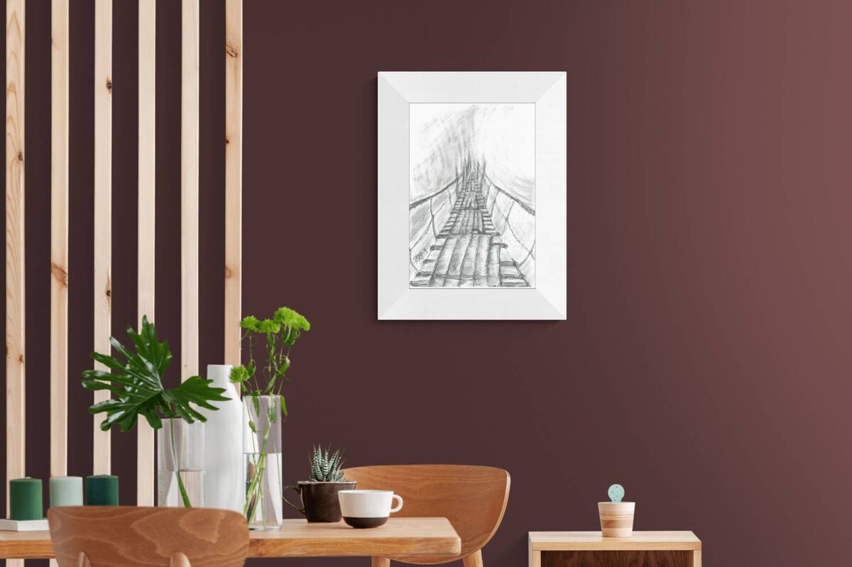 Original surreal charcoal drawing Toward the Unknown by Diana Dimova TRAXI, featuring a suspension bridge in mist exploring transition in black and white. A framed black and white sketch of a suspension bridge hanging over water, displayed on a dark brown wall in a modern room setting with wooden furniture and plants.