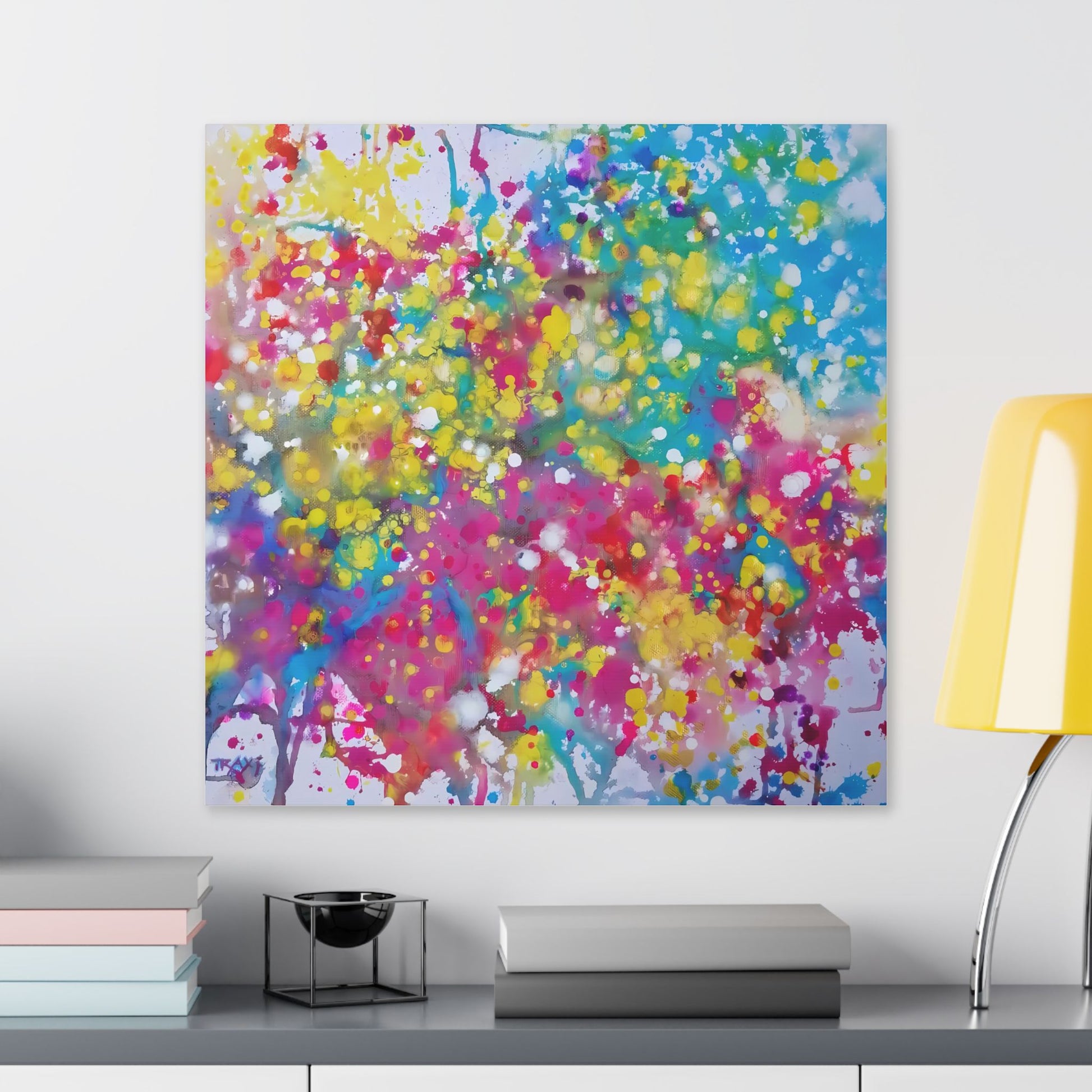Painting of happiness 2 vibrant abstract canvas print by award-winning contemporary artist Diana Dimova - TRAXI, featuring radiant pink, yellow, and blue colors. A square abstract wall art print featuring a vibrant splatter pattern in pink, yellow, blue, green, and purple, displayed on a light gray wall above a shelf with books and a yellow lamp. Painting of happiness 2 vibrant abstract canvas print by award-winning contemporary artist Diana Dimova - TRAXI, featuring radiant pink, yellow, and blue colors.