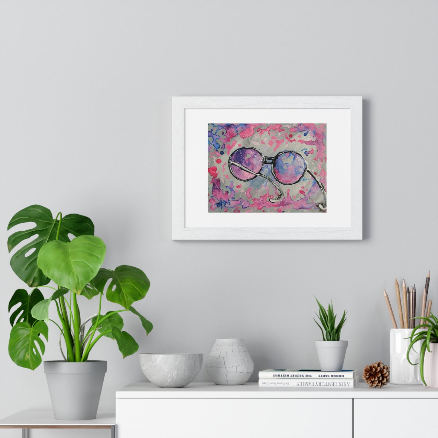 Pink glasses framed art print by contemporary artist TRAXI, featuring a joyful and optimistic theme on museum-quality archival matte paper. A framed abstract art print featuring purple and pink glasses on a textured pink background, displayed on a light gray wall above a white console table with plants and decorative items.