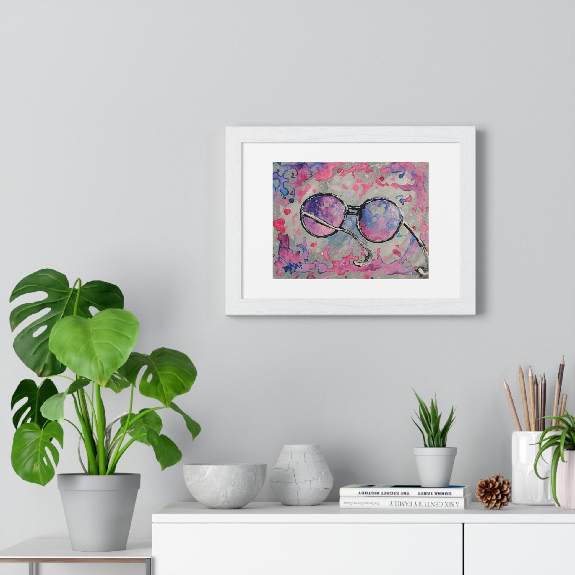Pink glasses framed art print by contemporary artist TRAXI, featuring a joyful and optimistic theme on museum-quality archival matte paper. A framed abstract art print featuring purple and pink glasses on a textured pink background, displayed on a light gray wall above a white console table with plants and decorative items.
