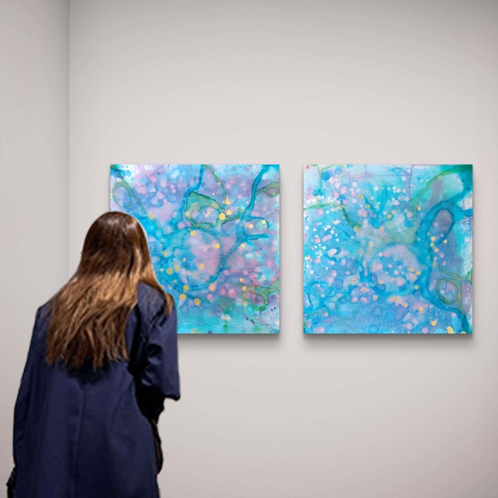 Original abstract diptych painting titled 'Rearrangement' by TRAXI, featuring textured layers in dialogue. The artwork explores themes of renewal, transition, and reconstruction through contemporary abstract expressionism. A woman with long brown hair, seen from behind, stands in an art gallery looking at two abstract square paintings hanging on a light gray wall. The paintings feature swirling patterns of blue, teal, purple, and pink with small yellow accents.
