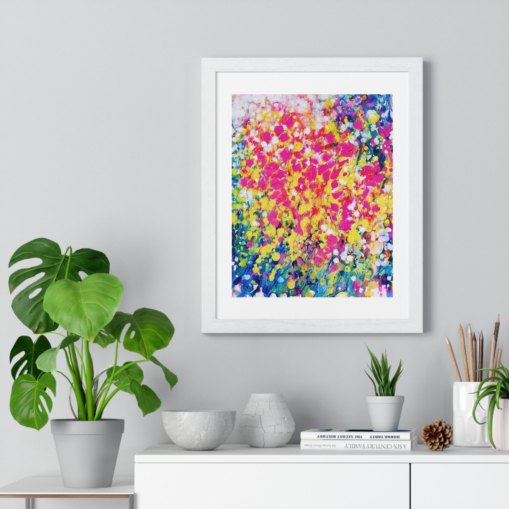 Summer flowers framed art print by contemporary artist TRAXI, featuring vibrant abstract floral colors on museum-quality archival matte paper. A framed abstract art print featuring a vibrant, textured paint pattern in pink, yellow, blue, and white, displayed on a light gray wall above a white console table with plants and decorative items.