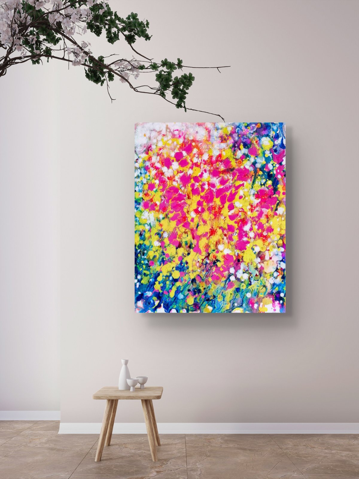 Summer flowers limited edition canvas print by contemporary artist TRAXI, featuring vibrant abstract fluid acrylic colors and warm seasonal energy in a 50x60 cm format. A colorful abstract canvas wall art print featuring vibrant splashes of pink, yellow, blue, and white, displayed on a light beige wall above a small wooden stool in a minimalist room setting.