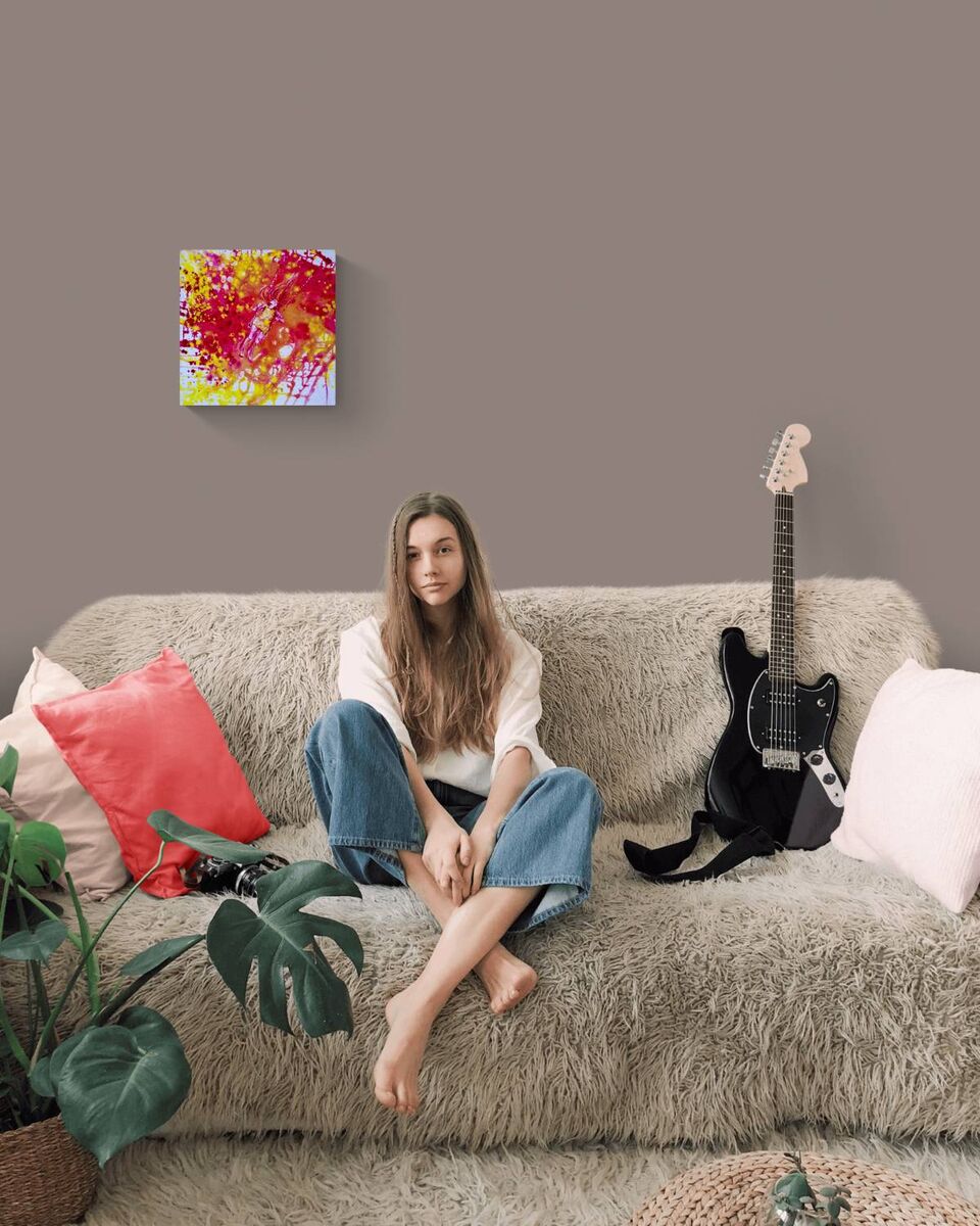 Sunshine Swing original fluid acrylic abstract painting by Diana Dimova TRAXI ART. Finalist Award winner 2022. A woman sits on a shaggy beige sofa in a styled living room setting, with a colorful abstract painting on the wall, a black electric guitar, and decorative pillows.