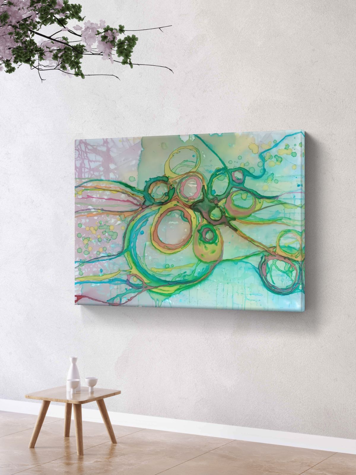 Original abstract painting titled 'The Balance' by TRAXI featuring serene textures and organic rhythms. The artwork represents the culmination of the series States of Becoming, exploring harmony, nature, and spiritual equilibrium. Abstract canvas wall art featuring flowing green, teal, yellow, and peach brushstrokes over a pale aqua and lavender background, displayed on a light textured wall above a small wooden side table.