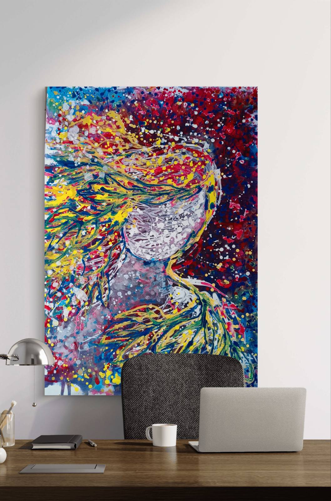 A colorful abstract canvas wall art print depicting a stylized female figure with flowing multicolored hair, displayed on a white wall above a wooden desk with a laptop, mug, and lamp. Autumn’s Smile original abstract portrait fluid acrylic painting with warm burgundy, gold, and orange tones representing feminine autumn energy by Diana Dimova TRAXI ART.