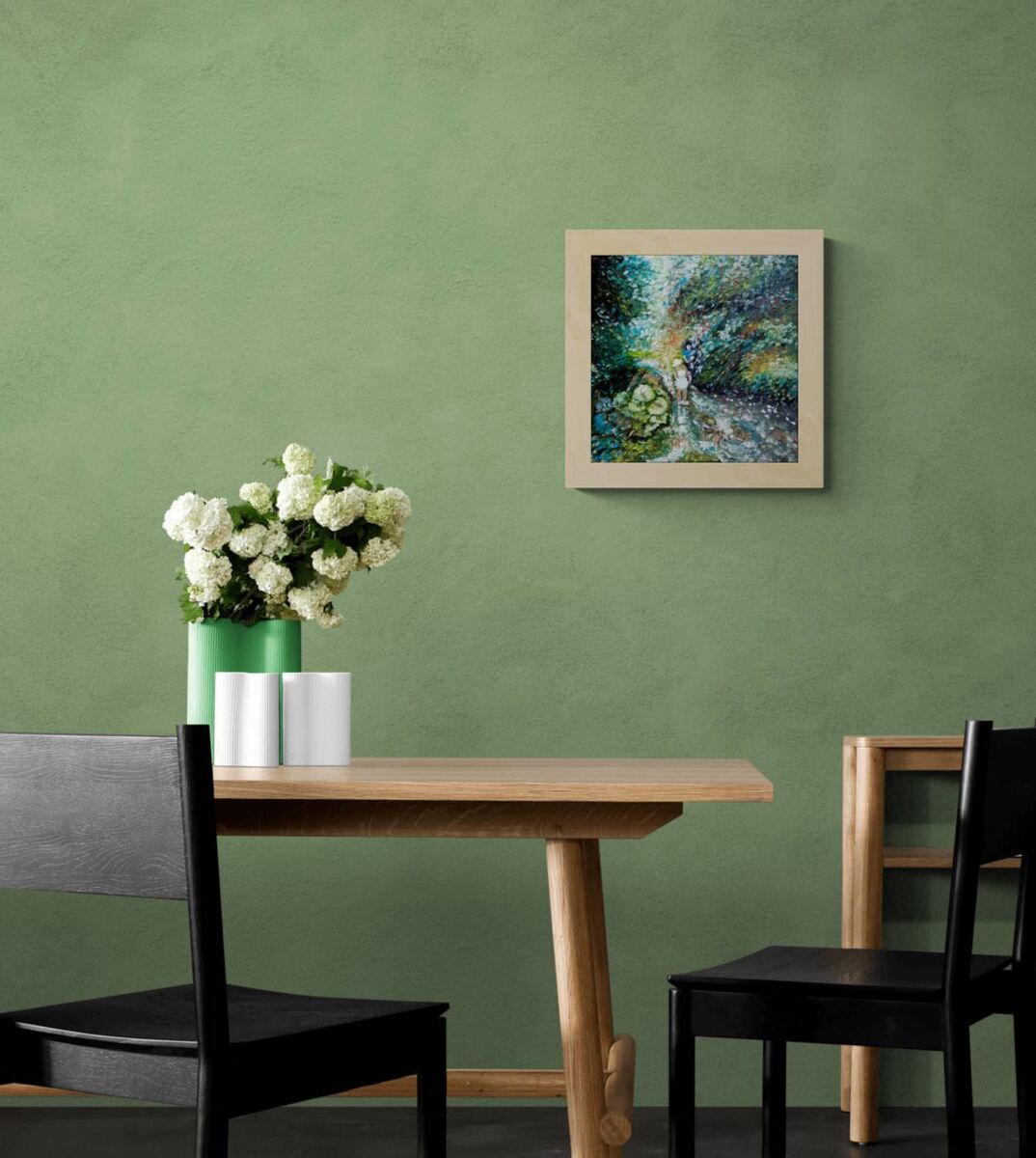A framed abstract landscape painting with blue, green, and white tones hangs on a textured sage green wall above a wooden dining table with black chairs and a vase of white flowers. By the River original impressionist oil painting with fresh green and white tones depicting a young girl by the river by Diana Dimova TRAXI ART.