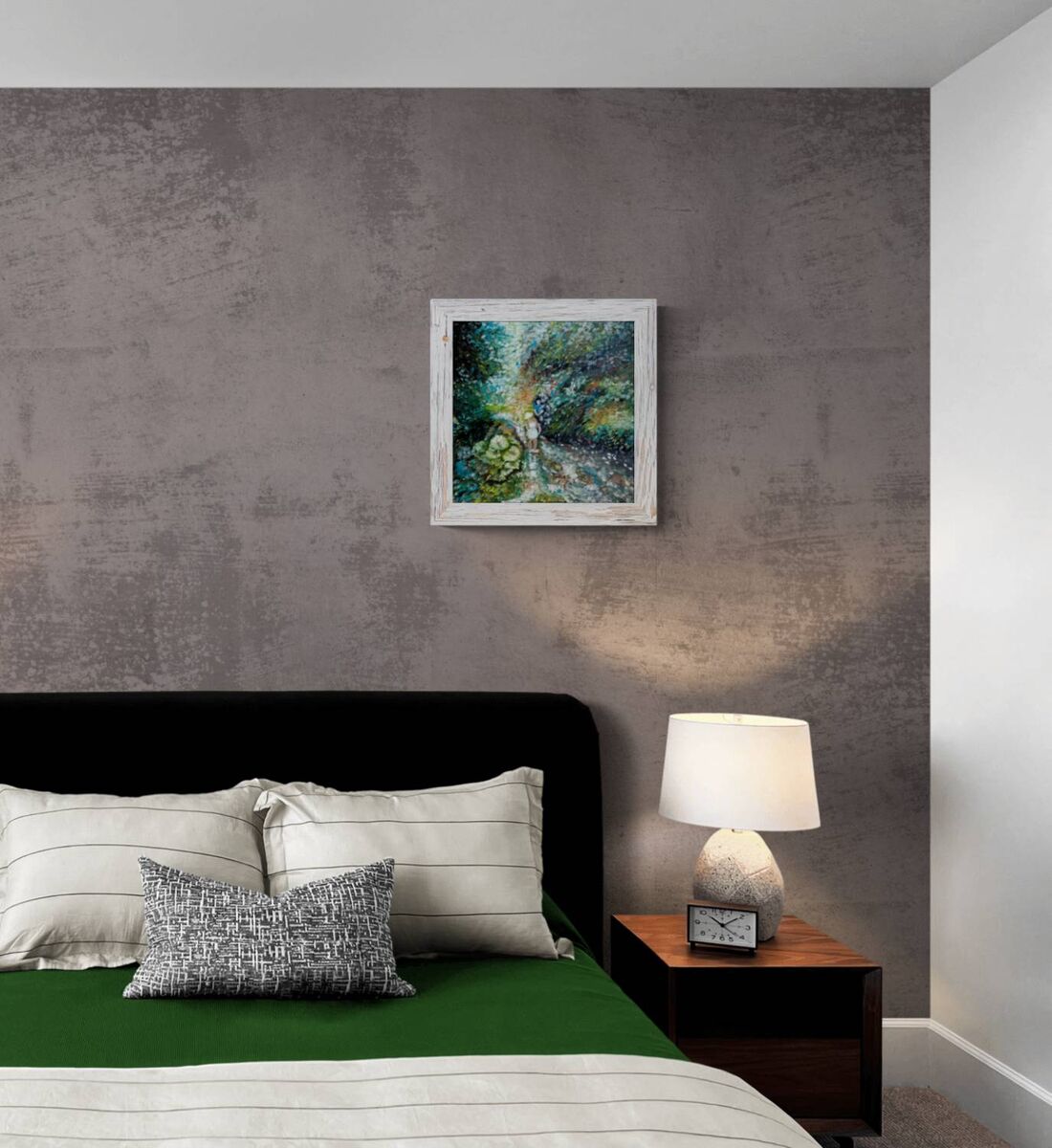 A framed landscape painting of a forest stream hangs on a textured gray wall above a bed with green bedding and striped pillows in a modern bedroom setting. By the River original impressionist oil painting with fresh green and white tones depicting a young girl by the river by Diana Dimova TRAXI ART.