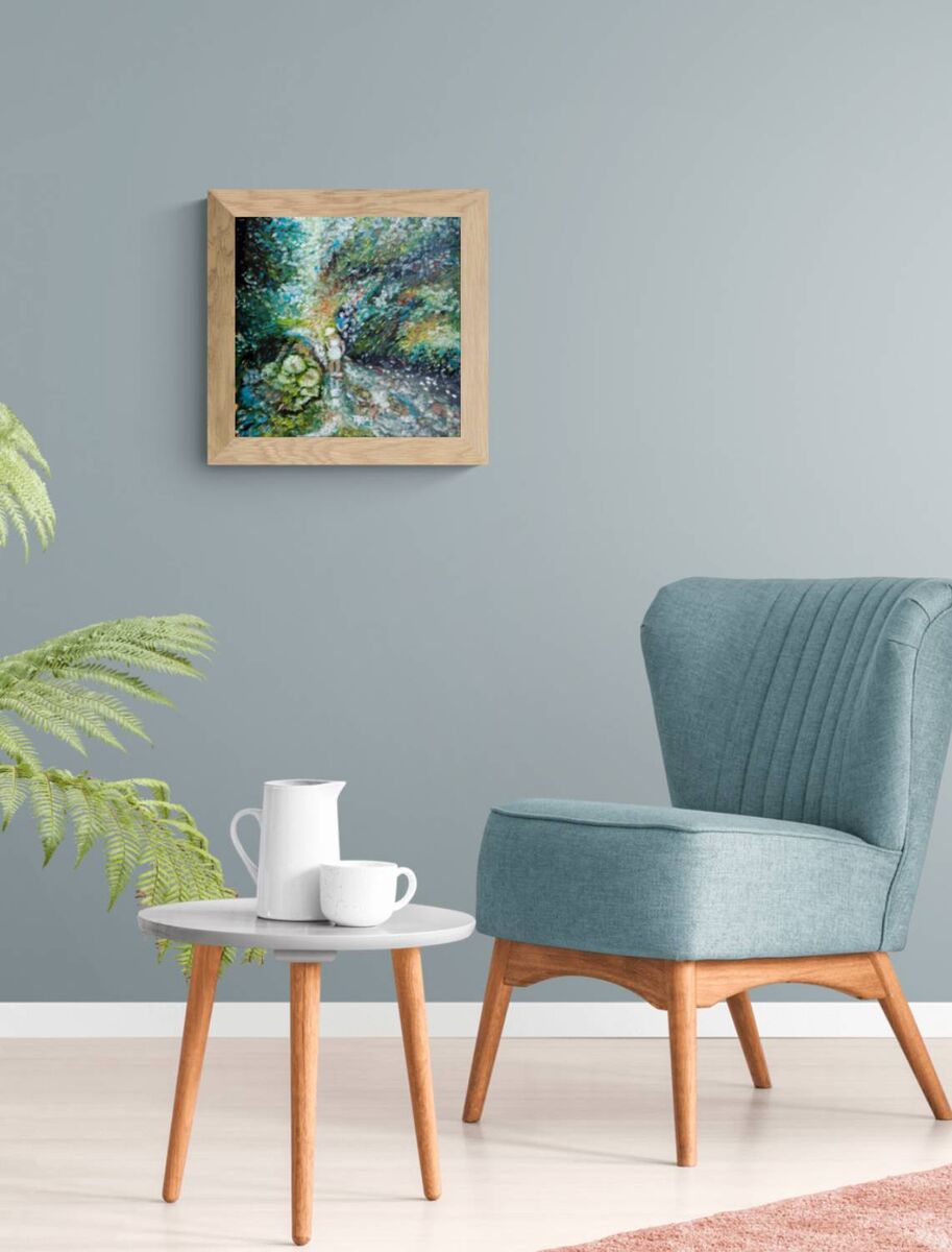 A framed abstract landscape painting featuring green, blue, and white tones, displayed on a gray wall in a modern living room setting with a teal armchair and a small side table. By the River original impressionist oil painting with fresh green and white tones depicting a young girl by the river by Diana Dimova TRAXI ART.