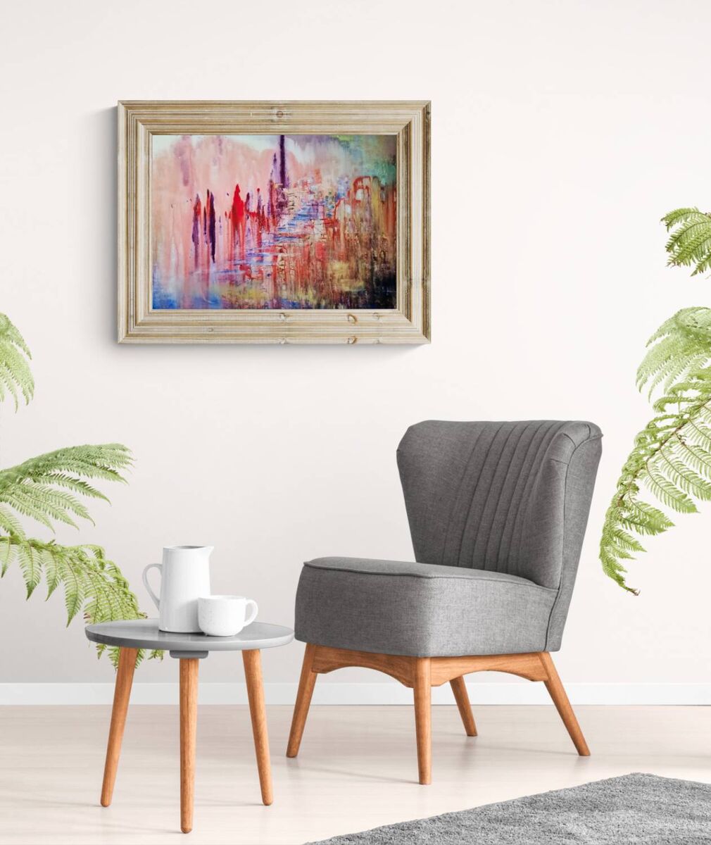 City of Memories original abstract lyrical fluid acrylic painting with dreamlike pink, violet, and orange tones representing nostalgia by Diana Dimova TRAXI ART. A framed abstract painting with vibrant red, blue, and pink brushstrokes hangs on a white wall above a gray upholstered armchair with wooden legs, next to a small round side table with a white mug and cup.