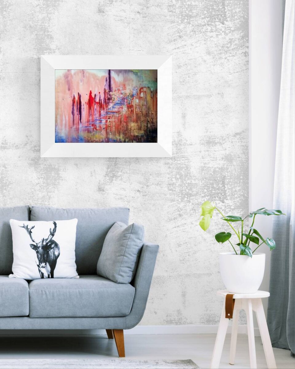 City of Memories limited edition premium horizontal canvas print with dreamlike pink, violet, and orange tones representing nostalgia by Diana Dimova TRAXI ART. A framed abstract painting with a white frame hangs on a textured light gray wall above a blue-gray sofa with decorative pillows. A small white stool with a potted green plant is on the right side of the room.