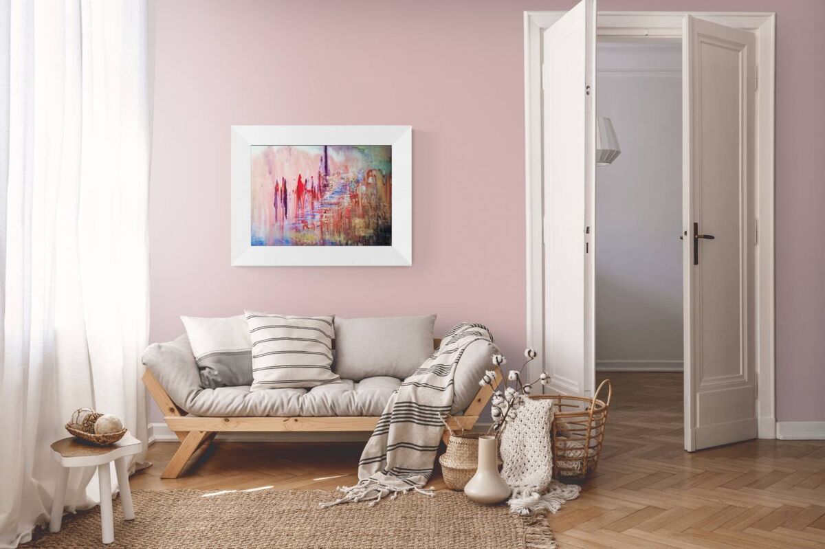 City of Memories original abstract lyrical fluid acrylic painting with dreamlike pink, violet, and orange tones representing nostalgia by Diana Dimova TRAXI ART. A framed abstract painting with vibrant red, blue, and yellow brushstrokes hangs on a dusty pink wall above a light wood daybed with gray cushions and a striped throw blanket in a bright, modern room.