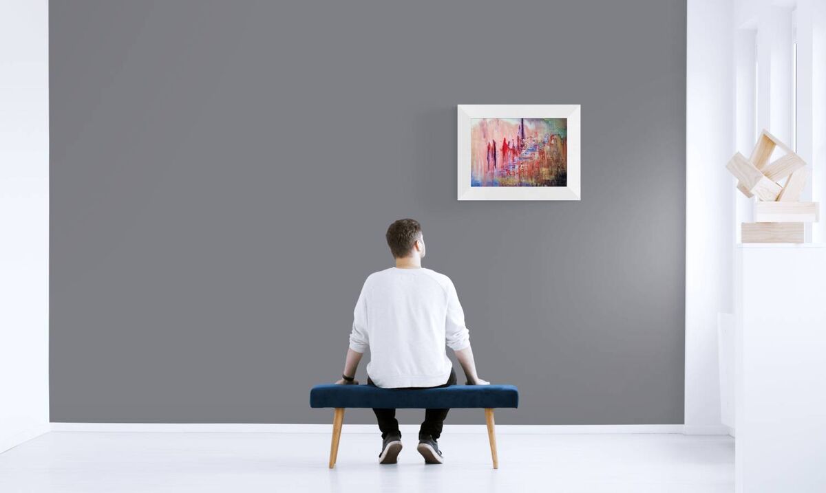 City of Memories original abstract lyrical fluid acrylic painting with dreamlike pink, violet, and orange tones representing nostalgia by Diana Dimova TRAXI ART. A man sits on a blue bench facing a gray wall, looking at a framed abstract painting. The scene is set in a minimalist white room with a sculptural wall lamp on the right.