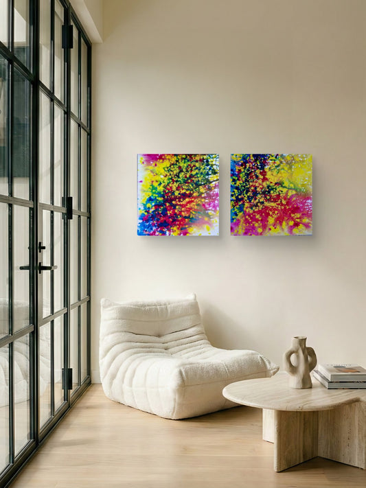 Fires Set original fluid acrylic abstract diptych in blue, red, orange, and yellow by Diana Dimova TRAXI ART. Finalist Award winner 2025. A pair of square abstract canvas prints with vibrant splatters of yellow, pink, blue, and green, displayed on a beige wall in a modern living room setting with a white lounge chair and a small wooden table.