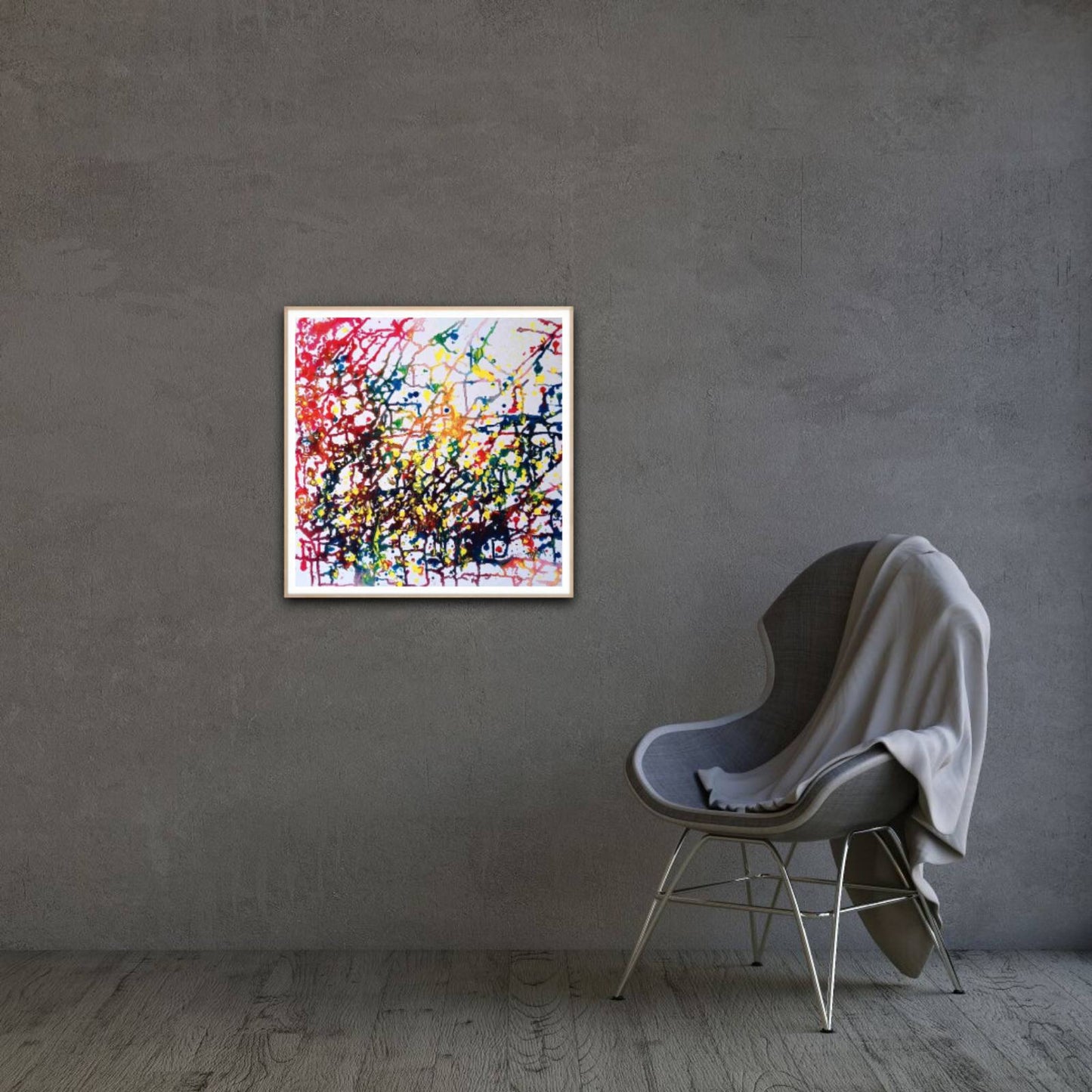 Flecks of Sunlight original fluid acrylic abstract painting by Diana Dimova TRAXI ART. Distinction Award winner 2025, San Francisco. A square framed abstract painting with vibrant splatters and drips in red, yellow, blue, green, and black on a white background, displayed on a textured gray wall next to a modern gray chair with a draped gray blanket.