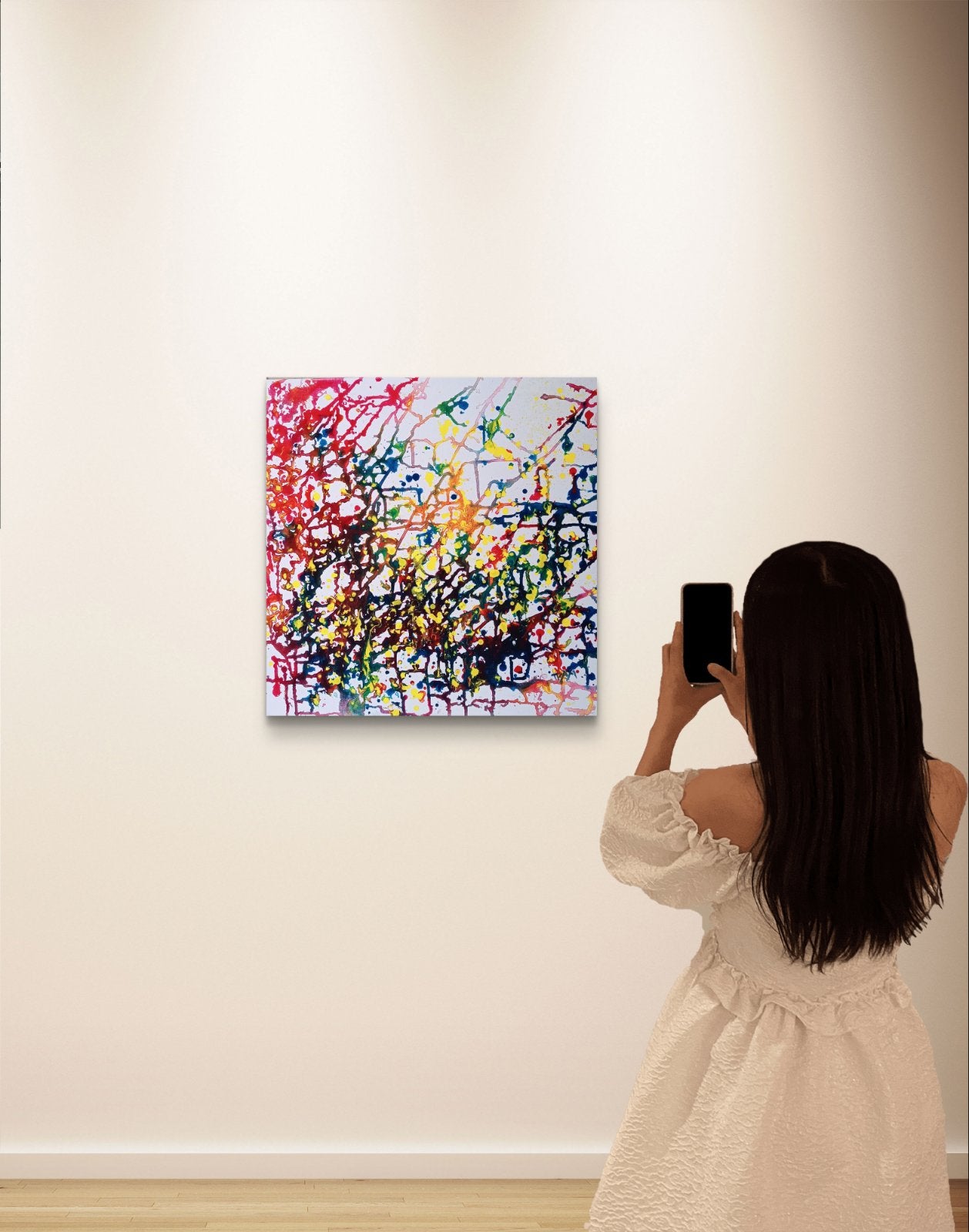 Flecks of Sunlight original fluid acrylic abstract painting by Diana Dimova TRAXI ART. Distinction Award winner 2025, San Francisco. A woman in a cream dress stands with her back to the camera, holding a smartphone to take a photo of a colorful abstract canvas painting hanging on a white gallery wall.
