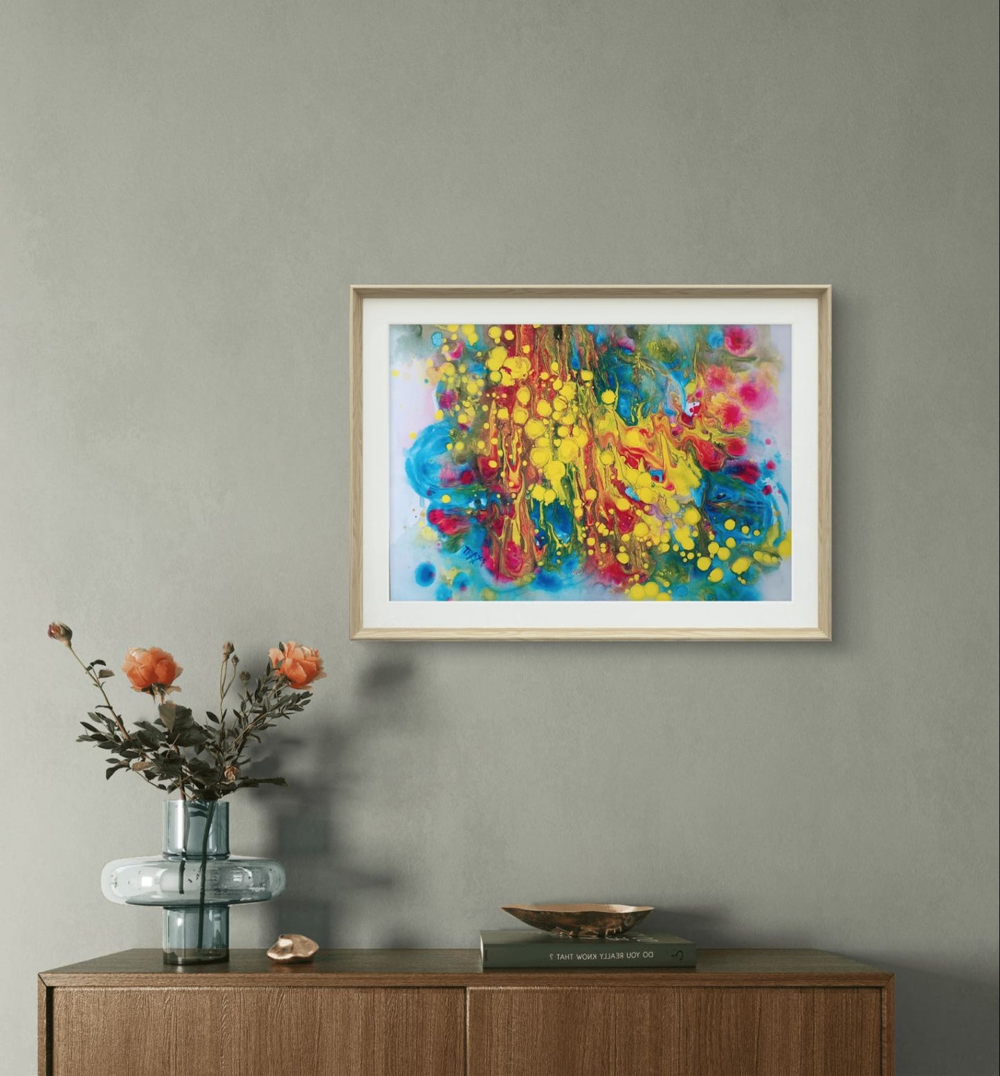 Fountains of Youth original watercolor abstract painting by Diana Dimova TRAXI ART, featuring dynamic yellow, blue, green, and red strokes. A framed abstract painting with vibrant splashes of blue, yellow, red, and pink hangs on a gray wall above a wooden console table with a vase of orange flowers and decorative objects.