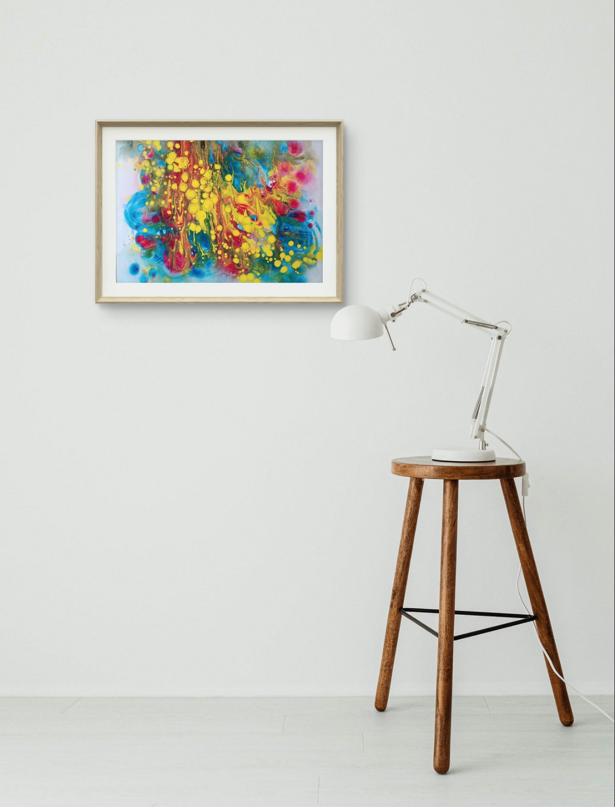 Fountains of Youth original watercolor abstract painting by Diana Dimova TRAXI ART, featuring dynamic yellow, blue, green, and red strokes. A colorful abstract painting in a light wood frame hangs on a white wall above a wooden stool with a white desk lamp.