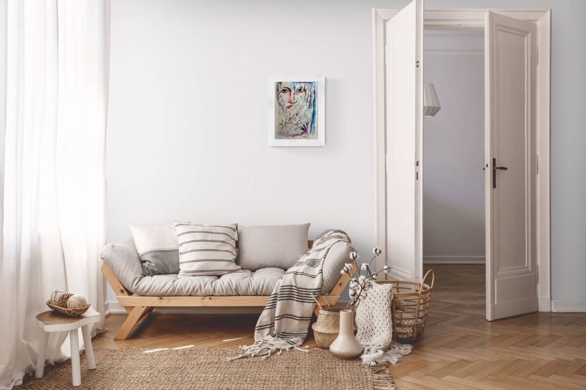 A light wood daybed with gray cushions and a striped throw blanket in a bright, minimalist living room. A small white side table, a woven rug, and a basket with decorative items sit in the foreground, with an open door and framed wall art in the background. Frozen original emotional watercolor portrait of a woman in blue and white tones by Diana Dimova TRAXI ART.