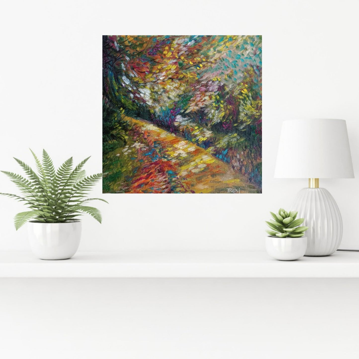 An impressionist-style square wall art print depicting a colorful forest path, displayed on a white shelf between a potted fern, a white lamp, and a small succulent. Impression in Autumn Colors original abstract impressionist oil painting with blue, yellow, orange, and red tones by Diana Dimova TRAXI ART.