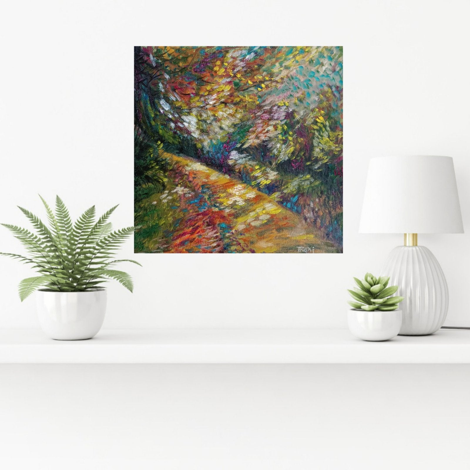 An impressionist-style square wall art print depicting a colorful forest path, displayed on a white shelf between a potted fern, a white lamp, and a small succulent. Impression in Autumn Colors original abstract impressionist oil painting with blue, yellow, orange, and red tones by Diana Dimova TRAXI ART.