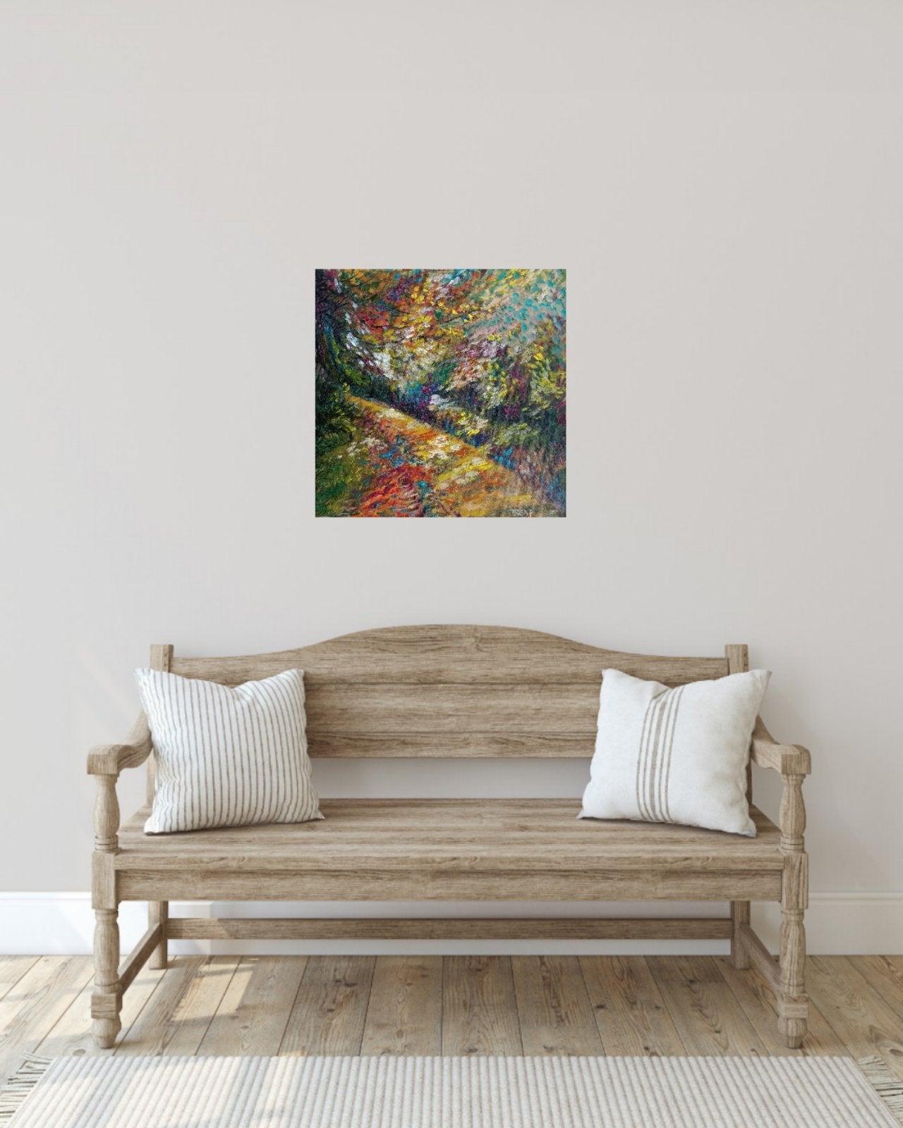 A square colorful painting of a forest path hangs on a light gray wall above a rustic wooden bench with two white striped pillows in a styled room setting. Impression in Autumn Colors original abstract impressionist oil painting with blue, yellow, orange, and red tones by Diana Dimova TRAXI ART.