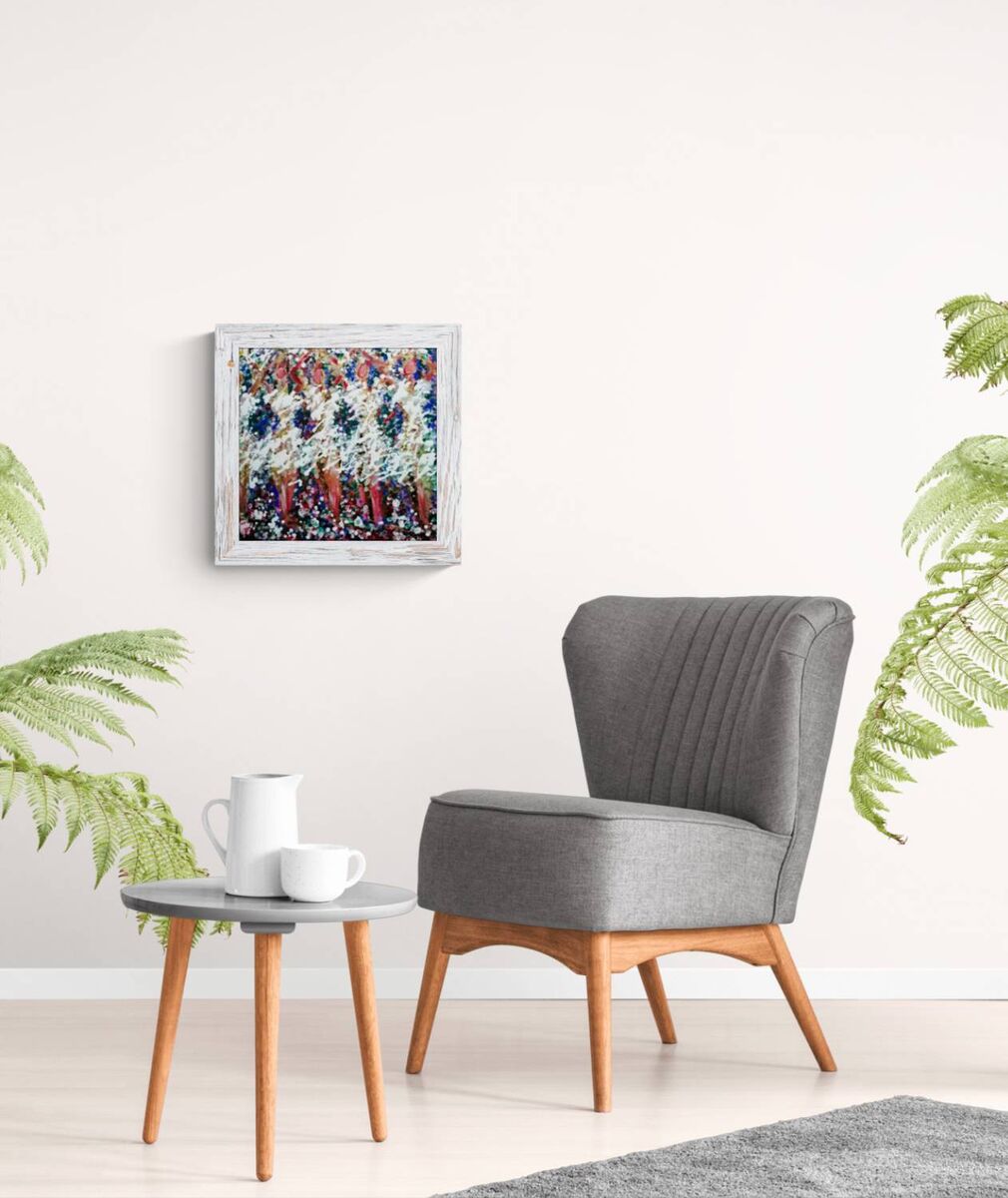 A framed abstract art print with a white frame hangs on a white wall above a gray upholstered armchair with wooden legs, next to a small round side table with a white mug and cup. Green plants frame the scene on both sides.In Spring the Trees Dance original abstract ballerina spring landscape fluid acrylic painting with deep burgundy, blue, and white tones representing plum trees in Bulgaria by Diana Dimova TRAXI ART.