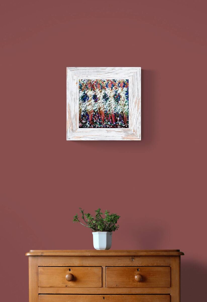 A framed abstract painting with a distressed white frame hangs on a dusty rose wall above a wooden chest of drawers with a small potted plant on top. In Spring the Trees Dance original abstract ballerina spring landscape fluid acrylic painting with deep burgundy, blue, and white tones representing plum trees in Bulgaria by Diana Dimova TRAXI ART.