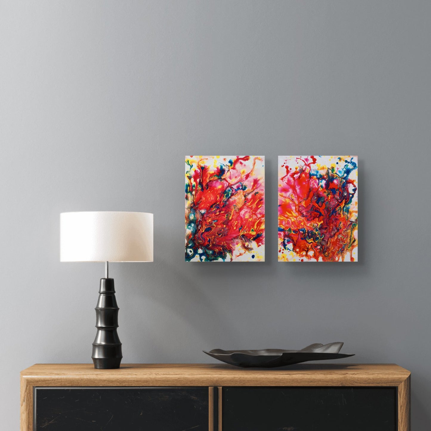 Magic Set original expressive red abstract diptych with fluid acrylic shapes by Diana Dimova TRAXI ART. Two square abstract canvas prints with vibrant red, orange, yellow, blue, and white paint splatters hang on a gray wall above a wooden console table with a black lamp and a black tray.