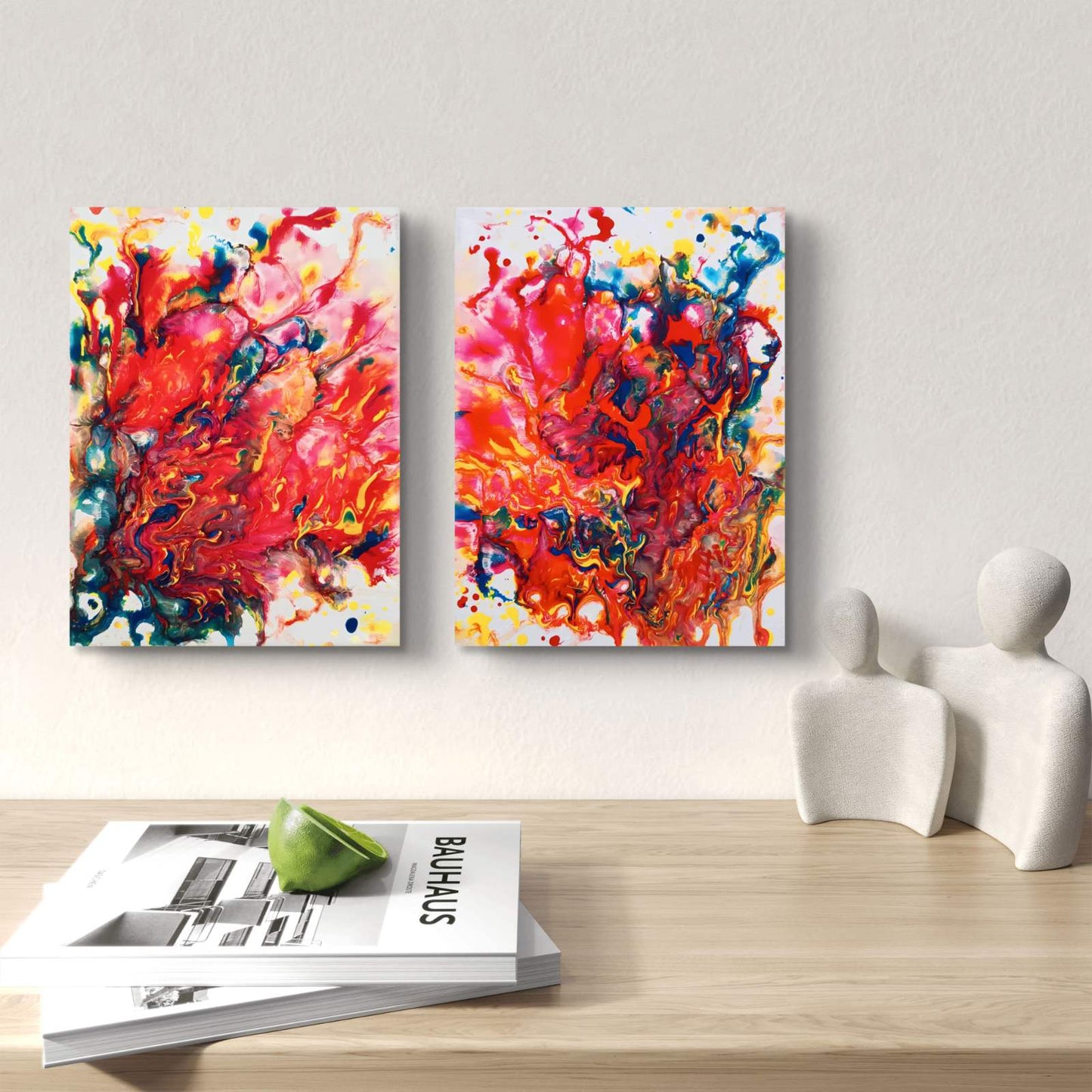 Magic Set original expressive red abstract diptych with fluid acrylic shapes by Diana Dimova TRAXI ART. Two vertical abstract canvas prints with vibrant red, orange, yellow, blue, and black paint splatters hang on a light gray wall above a wooden table with books and white decorative figures.