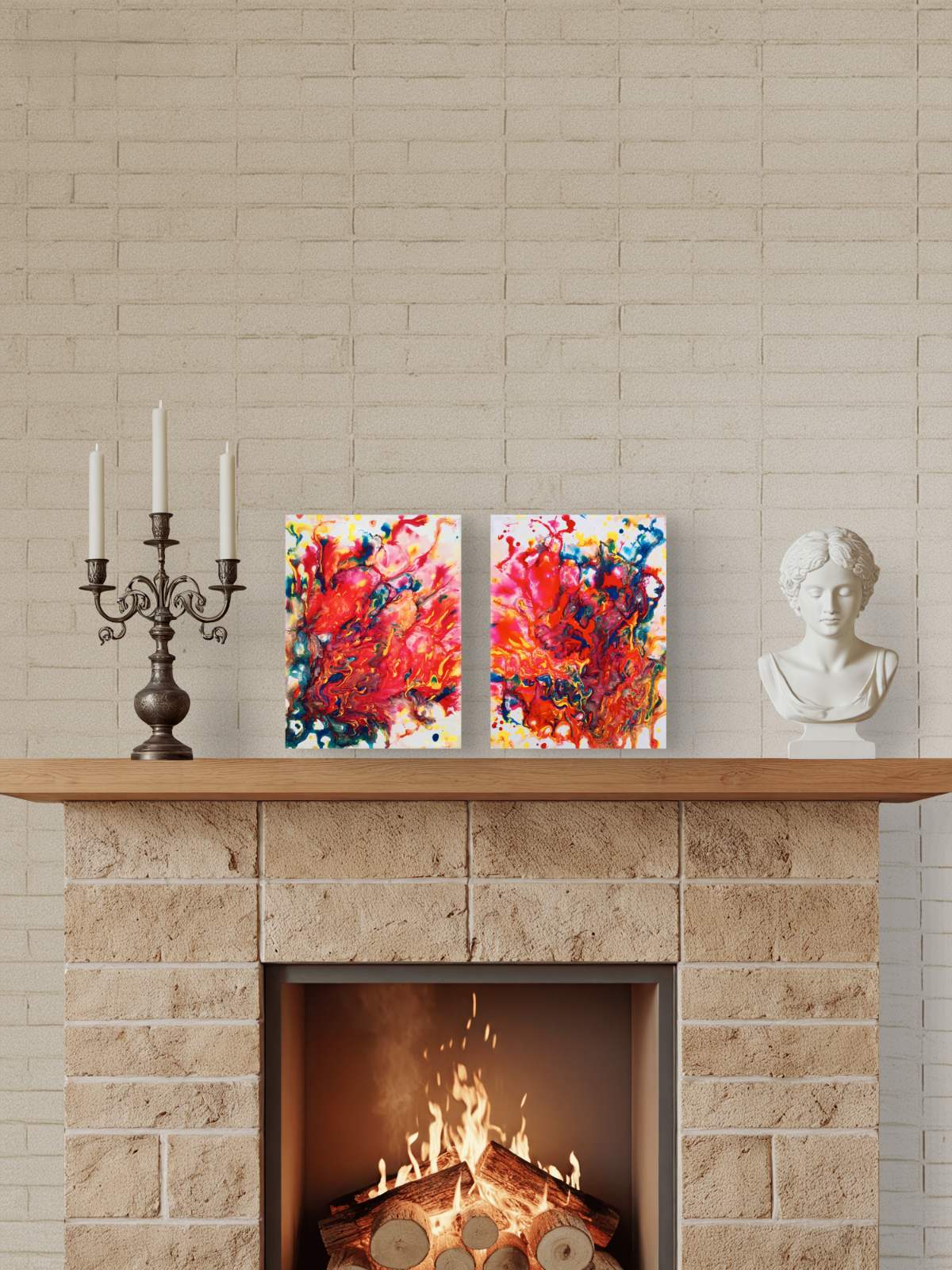 Magic Set original expressive red abstract diptych with fluid acrylic shapes by Diana Dimova TRAXI ART. Two square abstract canvas prints with vibrant red, orange, yellow, blue, and white brushstrokes are displayed on a wooden mantelpiece above a stone fireplace with a burning fire. Decorative items on the mantel include a black candelabra and a white classical bust, set against a light beige brick wall.
