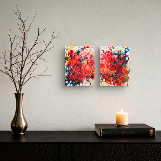 Magic Set original expressive red abstract diptych with fluid acrylic shapes by Diana Dimova TRAXI ART. Two square abstract canvas prints with vibrant red, orange, yellow, blue, and pink brushstrokes hang on a light gray wall above a dark wooden console table. A gold vase with bare branches sits on the left, and a lit candle on a book sits on the right.