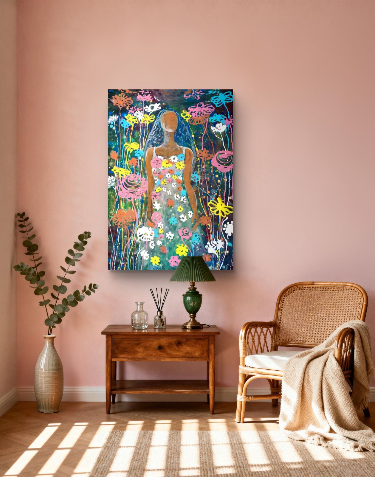 A colorful vertical canvas art print depicting a stylized figure surrounded by wildflowers, displayed on a dusty pink wall in a styled room setting with a wooden side table, lamp, rattan chair, and a vase of greenery. Palm Sunday original spring abstract fluid acrylic painting with vibrant floral colors celebrating feminine energy and Bulgarian heritage by Diana Dimova TRAXI ART.