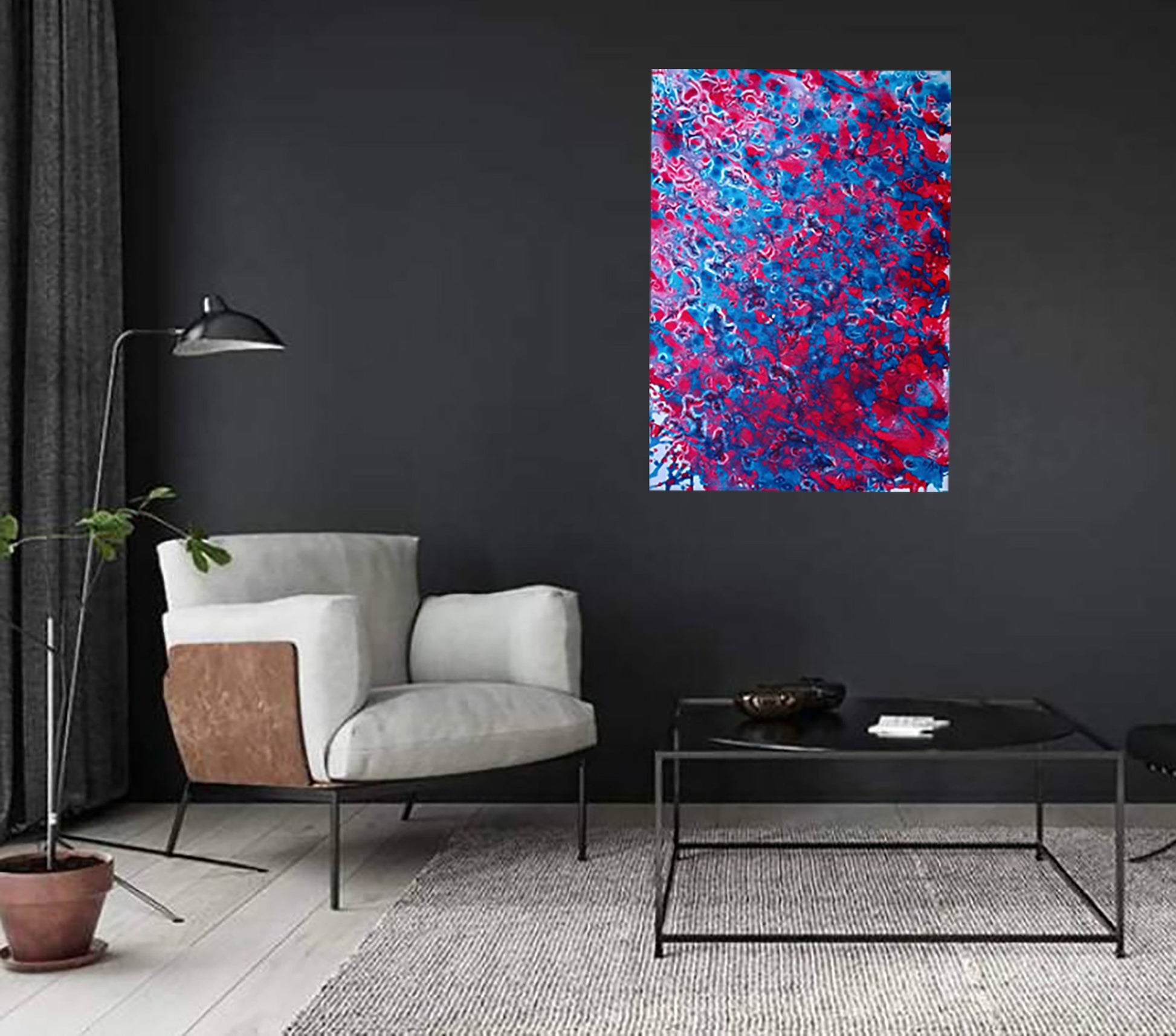 Red & Blue Balance original fluid acrylic abstract painting by Diana Dimova TRAXI ART. A colorful abstract canvas print with vibrant red and blue paint splatters hanging on a dark charcoal wall in a modern living room setting with a gray armchair, black coffee table, and floor lamp.