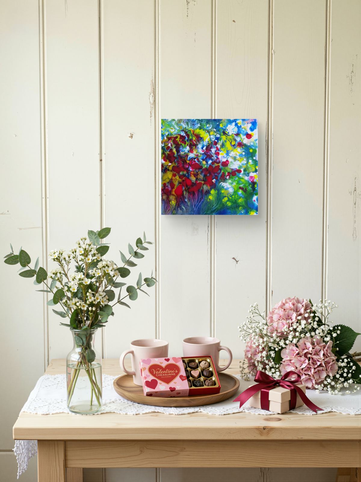 A colorful abstract painting hanging on a cream-colored wood-paneled wall above a light wood table styled with pink flowers, baby's breath, mugs, and a small gift box. Red Flowers original small abstract floral landscape fluid acrylic painting with vibrant red, green, and white tones by Diana Dimova TRAXI ART.