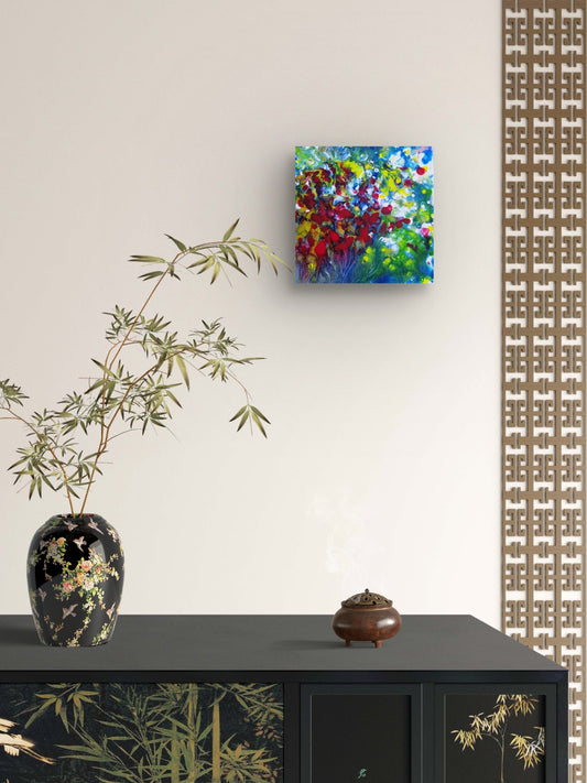 A colorful abstract canvas painting hanging on a beige wall above a dark cabinet with bamboo motifs, next to a black vase with green branches. Red Flowers original small abstract floral landscape fluid acrylic painting with vibrant red, green, and white tones by Diana Dimova TRAXI ART.