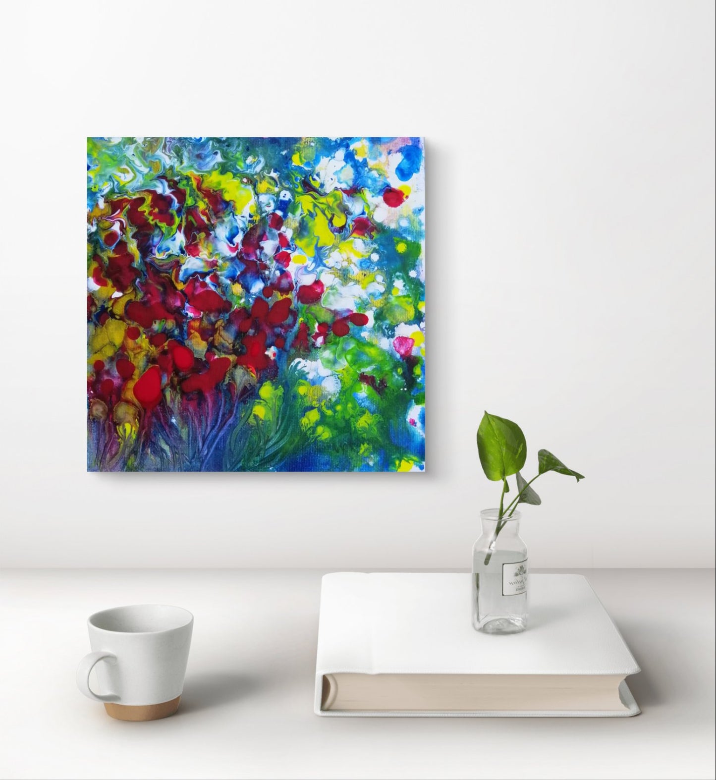 Colorful abstract painting on a white wall above a white surface with a cup, book, and plant. Red Flowers original small abstract floral landscape fluid acrylic painting with vibrant red, green, and white tones by Diana Dimova TRAXI ART.