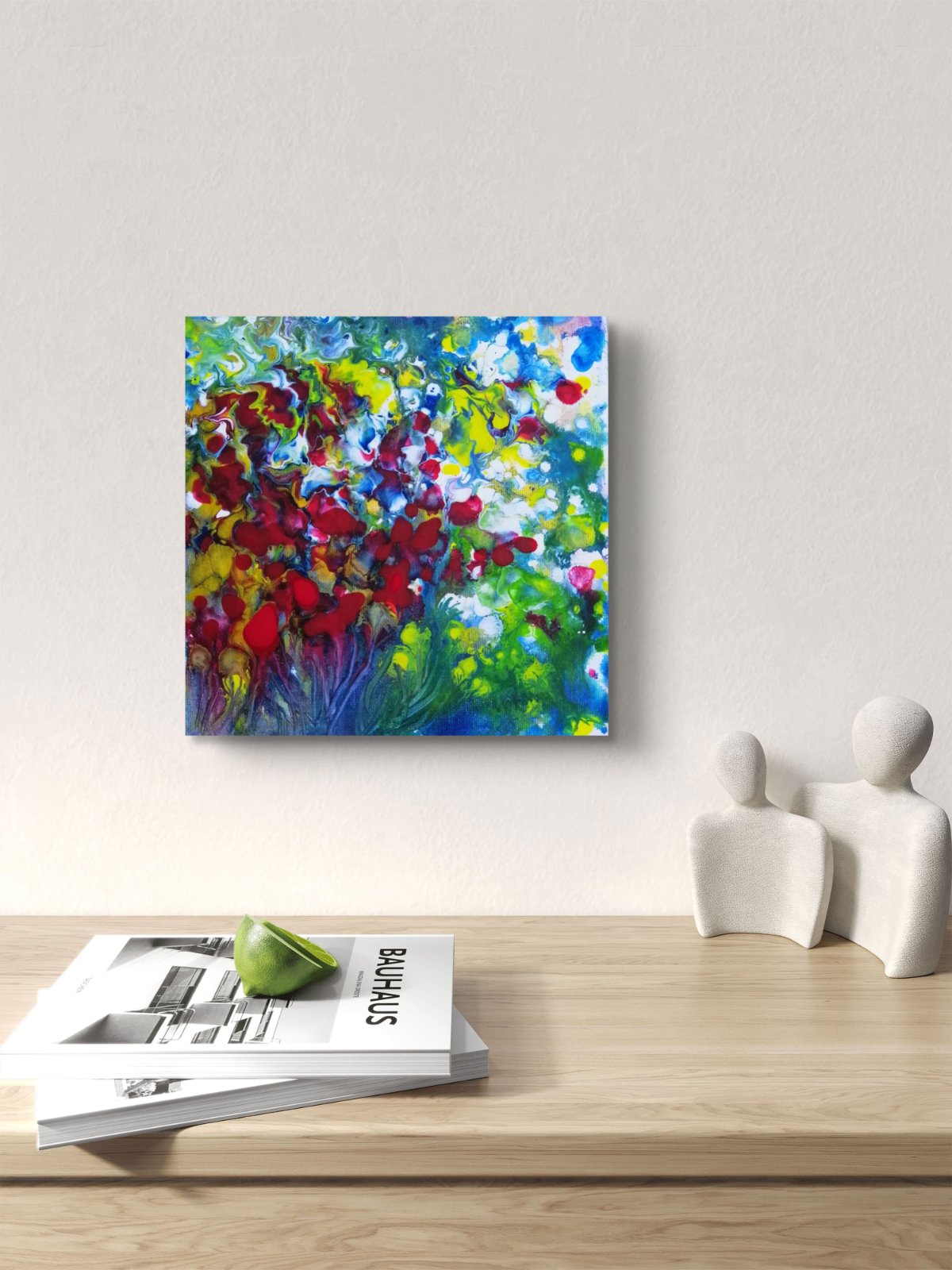 A colorful abstract canvas print featuring vibrant red, blue, green, yellow, and white brushstrokes, displayed on a light beige wall above a wooden shelf with books and white decorative figures. Red Flowers original small abstract floral landscape fluid acrylic painting with vibrant red, green, and white tones by Diana Dimova TRAXI ART.