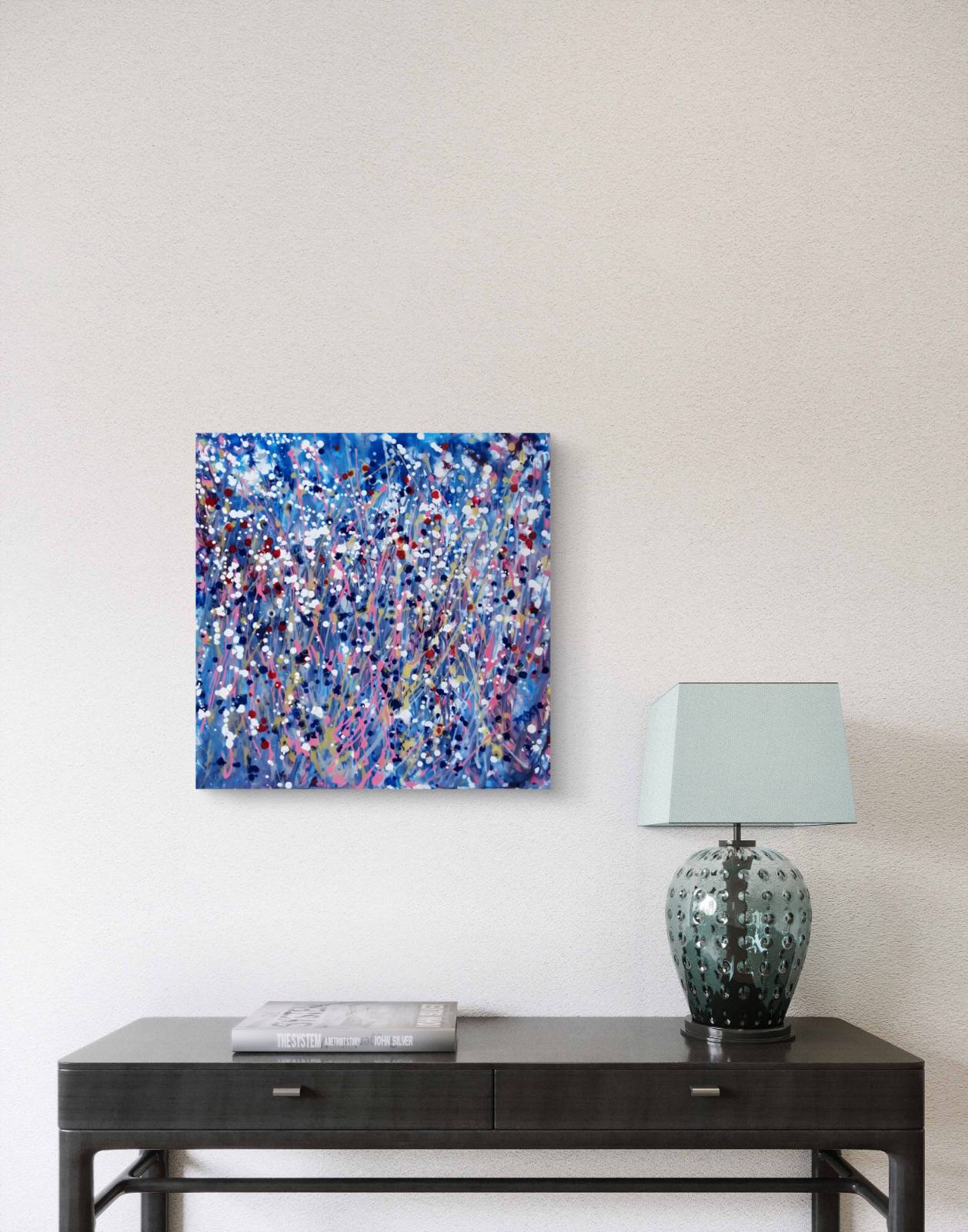 Starlight in the Grass limited edition premium square canvas print with calm blue, soft pink, and silver starlight tones by Diana Dimova TRAXI ART.  An abstract canvas painting with vibrant blue, purple, and white brushstrokes, displayed on a light beige wall above a dark wood console table with a lamp and a book.