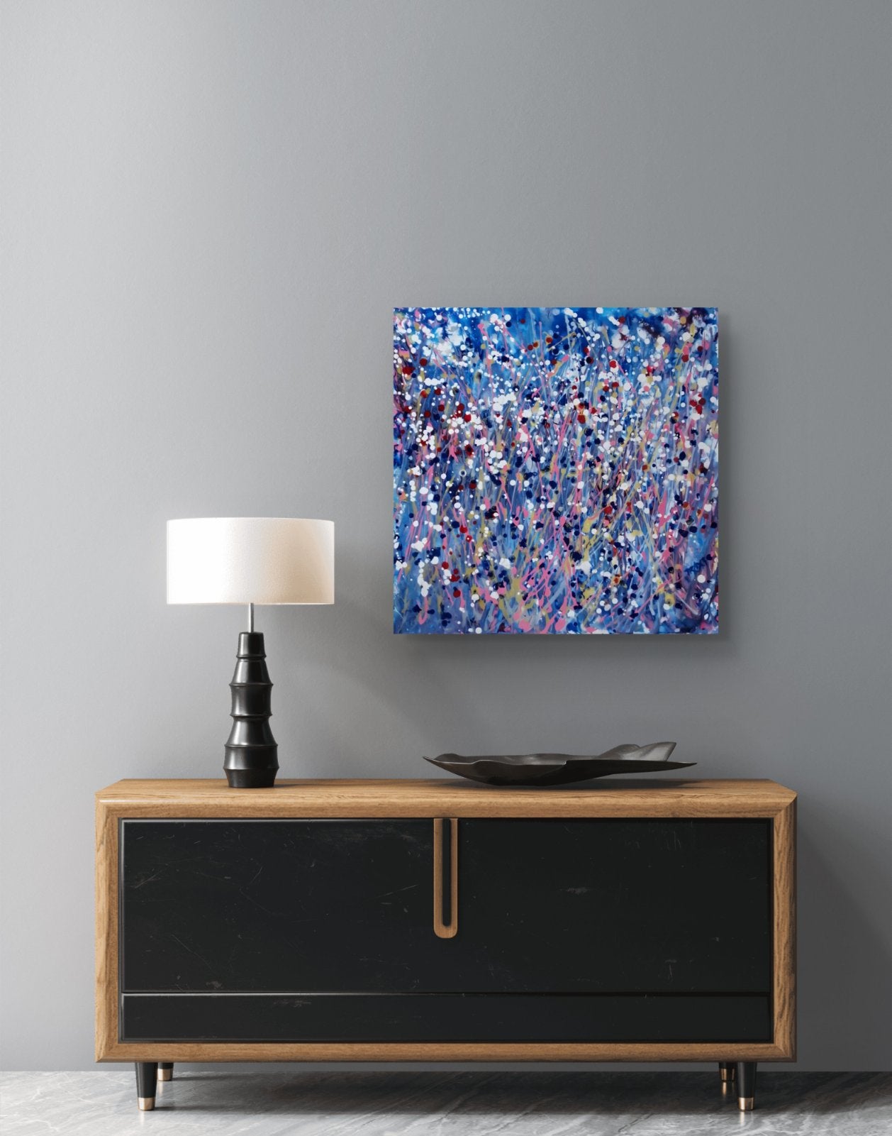 Starlight in the Grass original abstract square painting with calm blue, soft pink, and silver starlight tones by Diana Dimova TRAXI ART. An abstract canvas print featuring a vibrant blue, purple, and white paint splatter pattern, displayed on a gray wall above a mid-century modern sideboard with a black cabinet, wood frame, and a black table lamp.