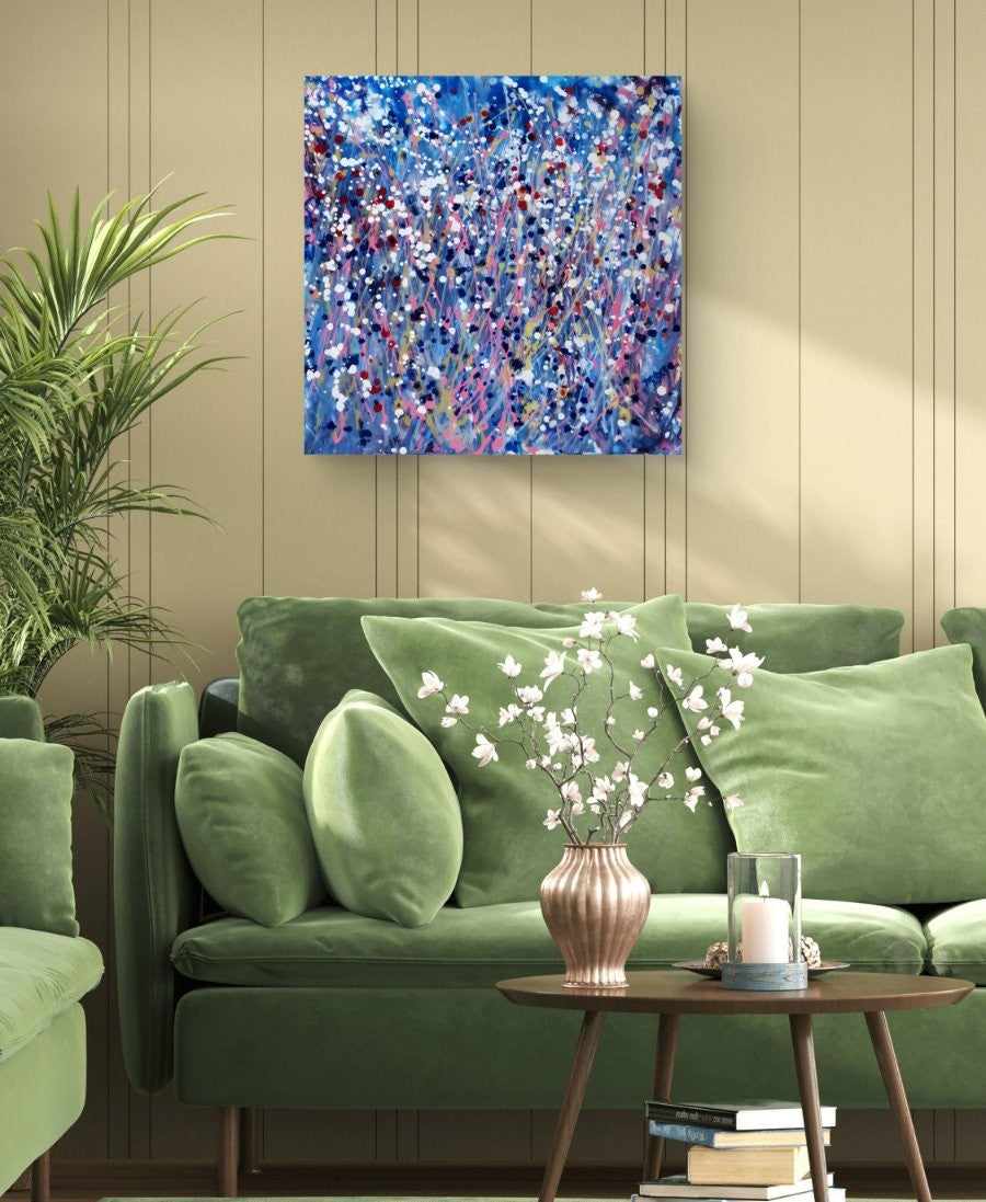 Starlight in the Grass original abstract square painting with calm blue, soft pink, and silver starlight tones by Diana Dimova TRAXI ART. An abstract canvas wall art print with a vibrant blue, purple, and white paint splatter design, displayed on a beige paneled wall above a green velvet sofa in a living room setting.