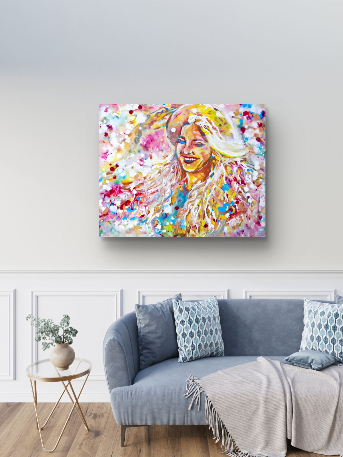 A colorful abstract portrait canvas print of a smiling woman with blonde hair, displayed on a light gray wall above a blue-gray sofa in a living room setting. Sun in the Hair limited edition premium horizontal canvas print depicting a smiling woman in a straw hat with pink and yellow summer light by Diana Dimova TRAXI ART.