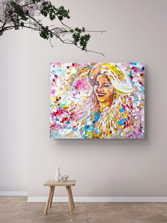 A colorful abstract portrait canvas print of a smiling woman, displayed on a light beige wall above a small wooden stool in a styled room  Sun in the Hair limited edition premium horizontal canvas print depicting a smiling woman in a straw hat with pink and yellow summer light by Diana Dimova TRAXI ART.setting.