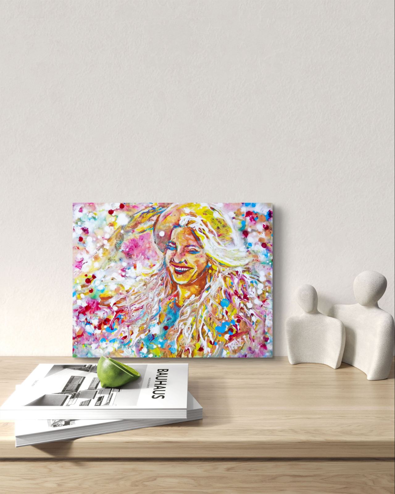A colorful abstract portrait canvas print leaning on a light wood shelf next to two white decorative figurines and a stack of magazines. Sun in the Hair original abstract feminine portrait fluid acrylic painting depicting a smiling woman in a straw hat with pink and yellow summer light by Diana Dimova TRAXI ART.