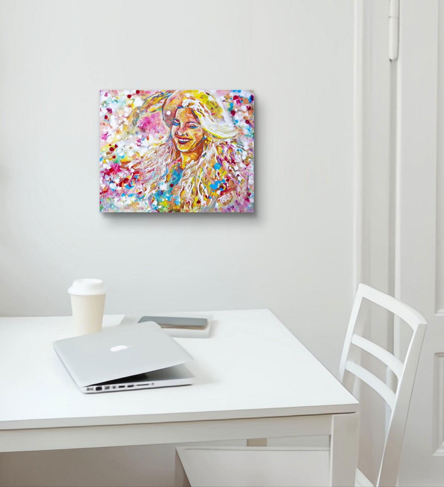 A colorful abstract portrait canvas print hanging on a white wall above a white desk with a laptop, coffee cup, and chair. Sun in the Hair original abstract feminine portrait fluid acrylic painting depicting a smiling woman in a straw hat with pink and yellow summer light by Diana Dimova TRAXI ART.