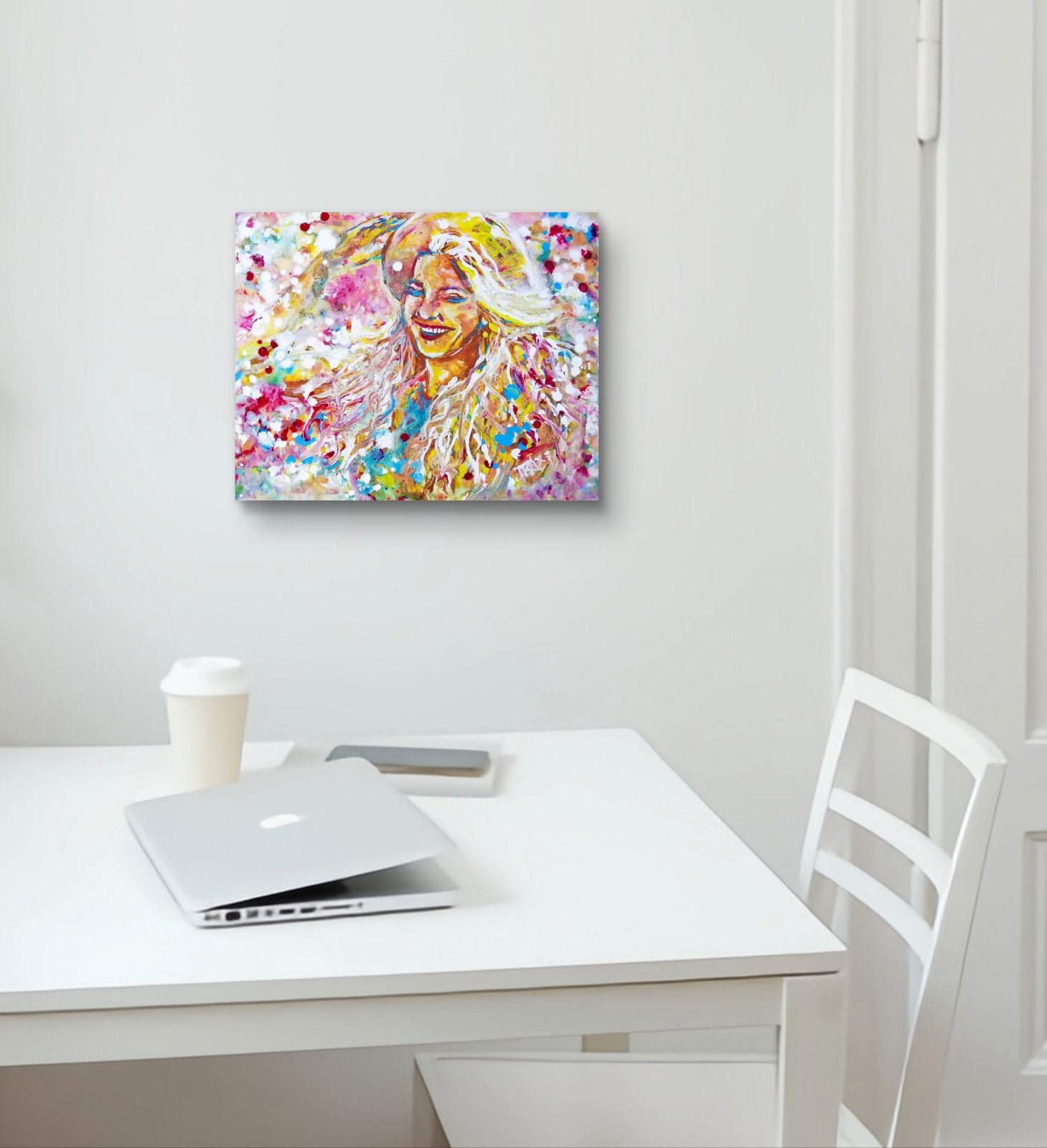 A colorful abstract portrait canvas print hanging on a white wall above a white desk with a laptop, coffee cup, and chair. Sun in the Hair original abstract feminine portrait fluid acrylic painting depicting a smiling woman in a straw hat with pink and yellow summer light by Diana Dimova TRAXI ART.