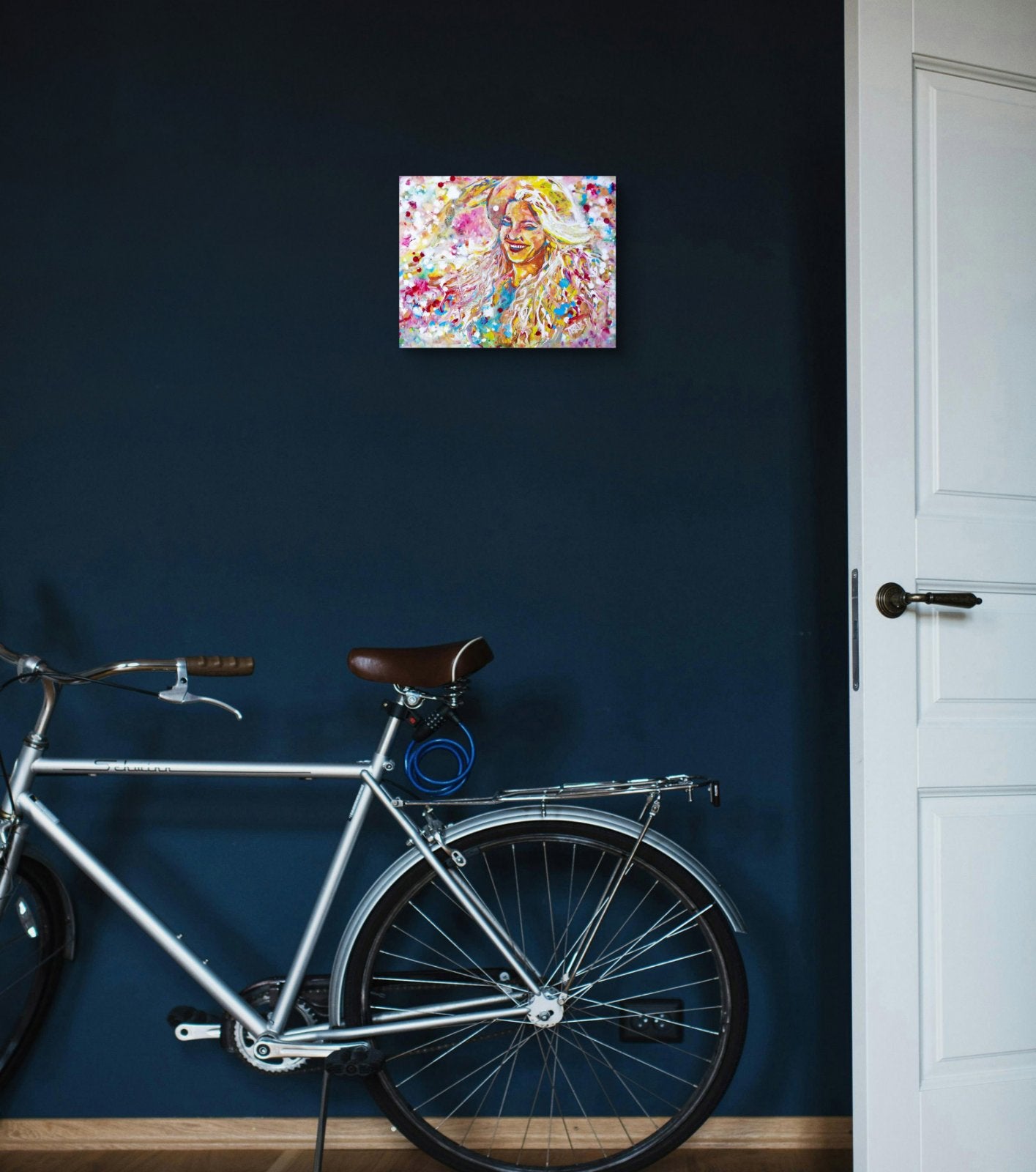 A colorful abstract painting of a woman with flowing hair hangs on a dark navy blue wall above a silver bicycle, next to a white door. Sun in the Hair original abstract feminine portrait fluid acrylic painting depicting a smiling woman in a straw hat with pink and yellow summer light by Diana Dimova TRAXI ART.