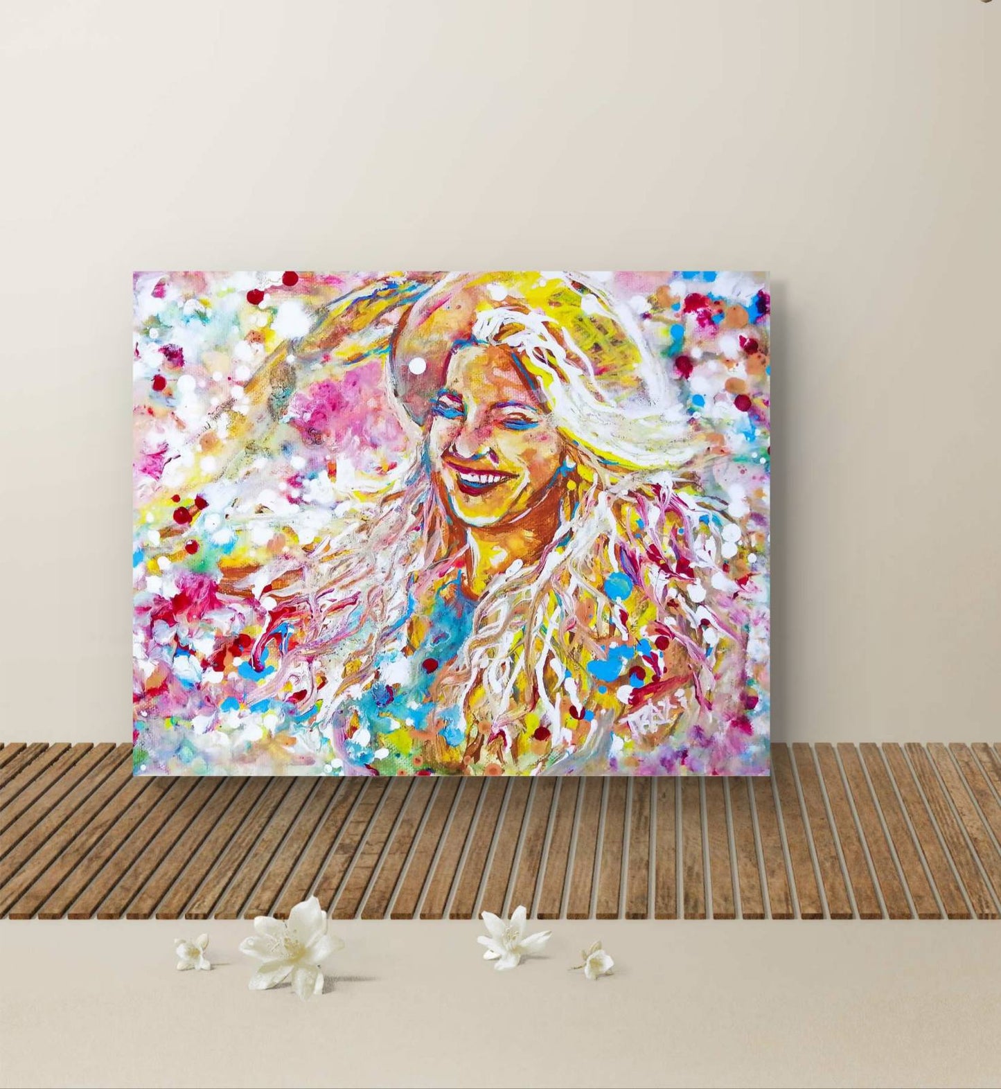 A colorful abstract portrait canvas print of a smiling woman with blonde hair, displayed on a wooden slatted surface against a light beige wall with small white flowers in the foreground. Sun in the Hair original abstract feminine portrait fluid acrylic painting depicting a smiling woman in a straw hat with pink and yellow summer light by Diana Dimova TRAXI ART.