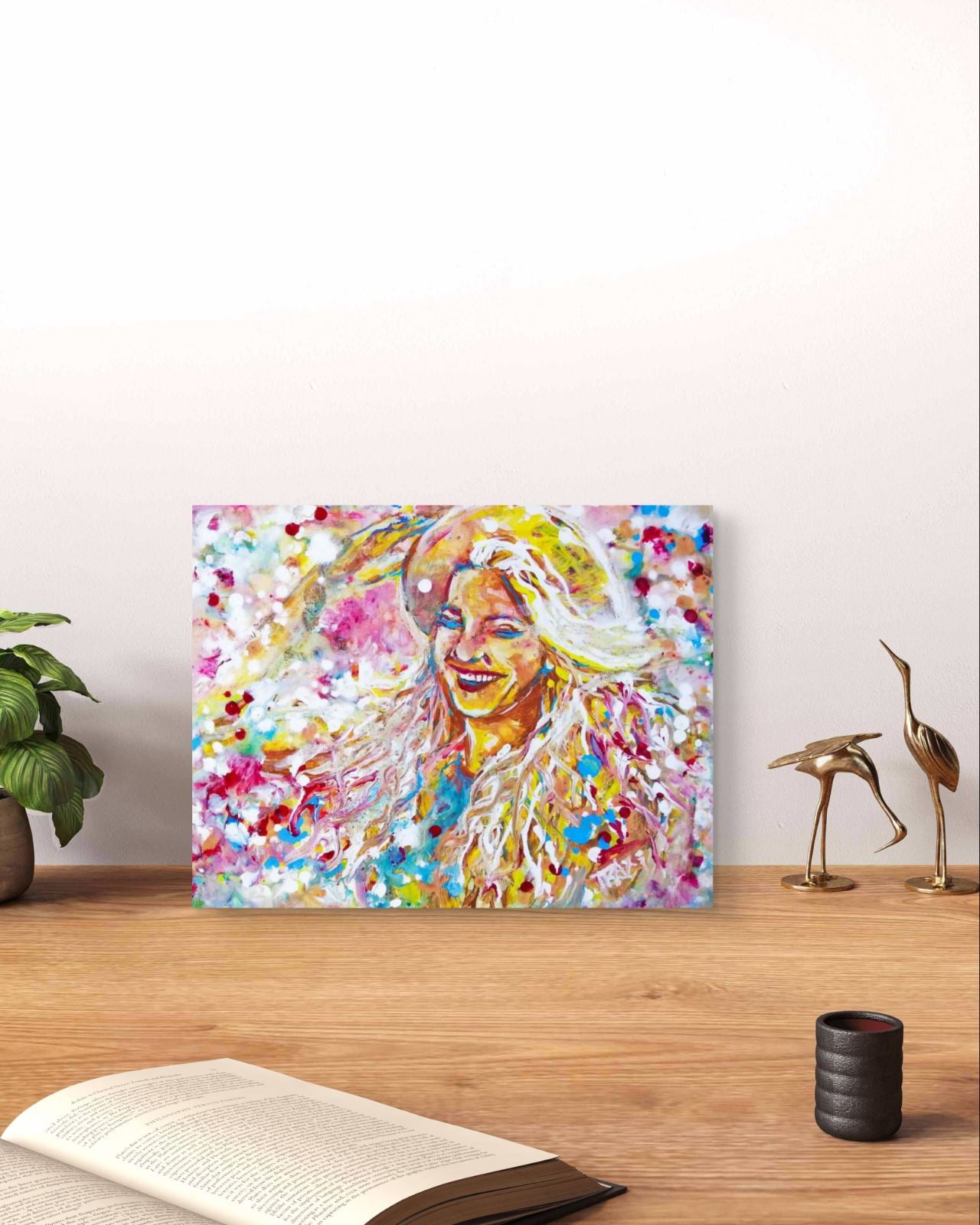 A colorful abstract portrait canvas print of a smiling woman with flowing hair, displayed on a wooden table in a styled room setting with a plant, book, and decorative cranes. Sun in the Hair original abstract feminine portrait fluid acrylic painting depicting a smiling woman in a straw hat with pink and yellow summer light by Diana Dimova TRAXI ART.